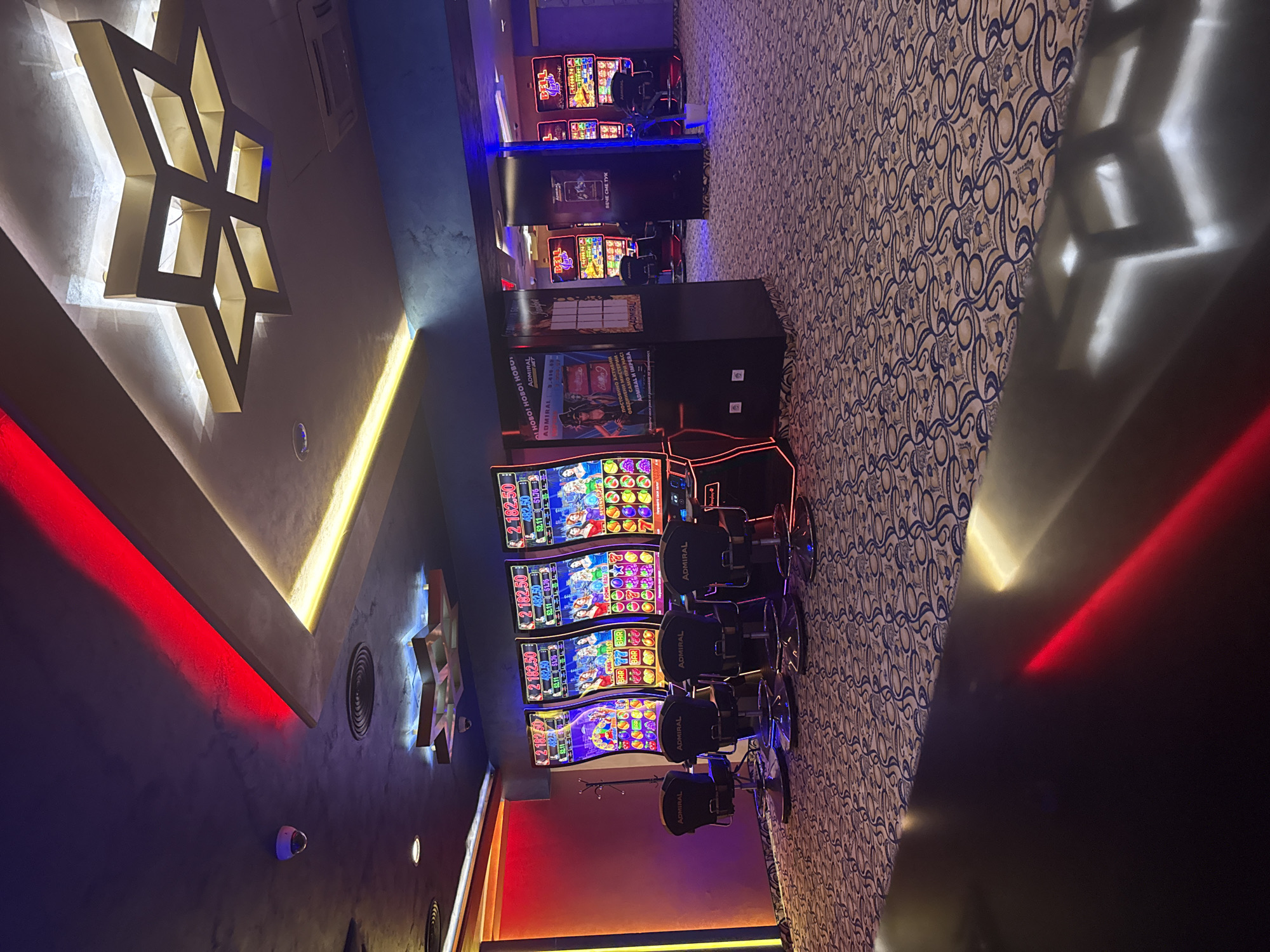 The image shows the interior of a casino in Pleven, Bulgaria. The scene includes rows of slot machines with chairs in front of them. The floor is covered with a patterned carpet. The walls have decorative lighting, including neon lights and a large star-shaped fixture. There are security cameras visible on the walls. Information displays are placed next to the slot machines. OCR identifies text on the machines and displays indicating currency amounts, phrases like "ADMIRAL," and phrases in Bulgarian.