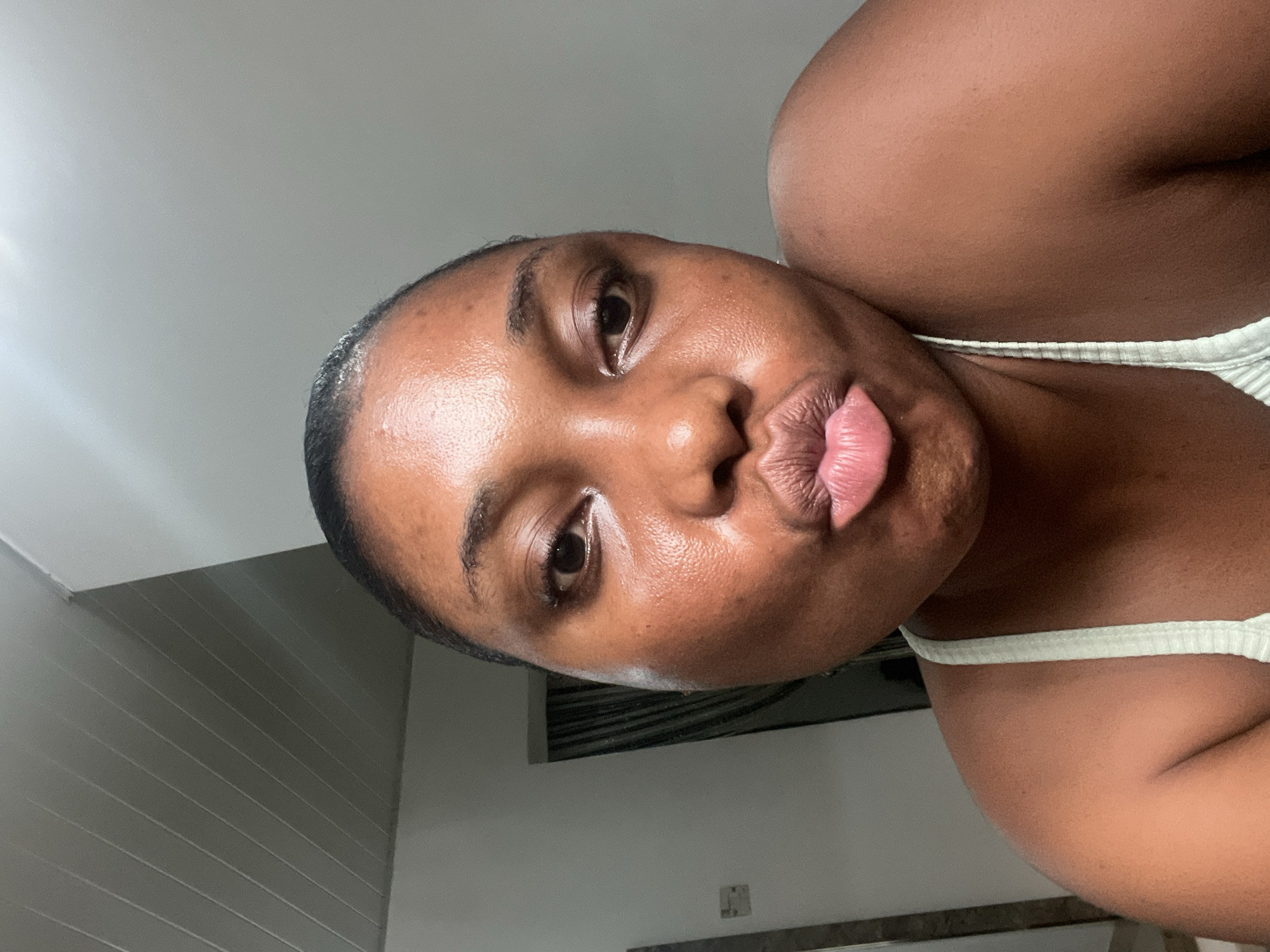 A dark-skinned person with slicked-back hair is posing indoors, with their lips puckered. They are wearing a white ribbed tank top. The location appears to be indoors, with a wall and ceiling visible in the background. The exact location within Abuja, Nigeria, cannot be determined from the image.