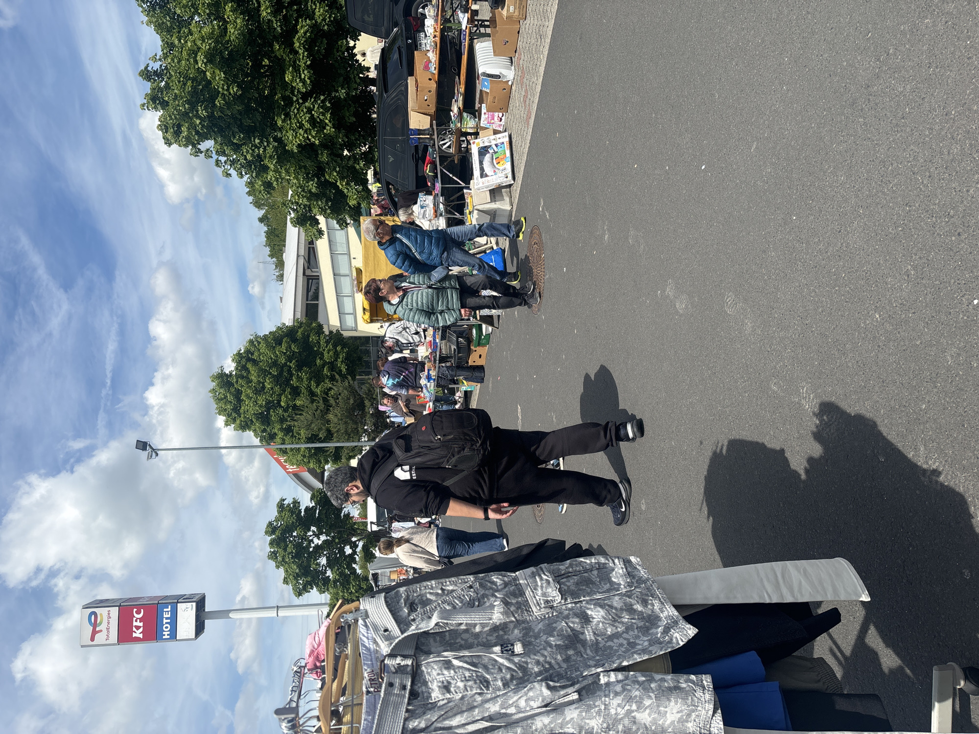This image depicts an outdoor flea market scene in Eichenzell, Germany. The market appears to be set up along a street. People are browsing items displayed on tables and racks. The foreground features a rack filled with clothes. A sign with TotalEnergies, KFC, and Hotel logos is visible. Trees line the street. The sky is blue with scattered clouds. There are people standing and walking among the stalls and cars parked nearby, suggesting vendors and visitors are engaging in commerce.