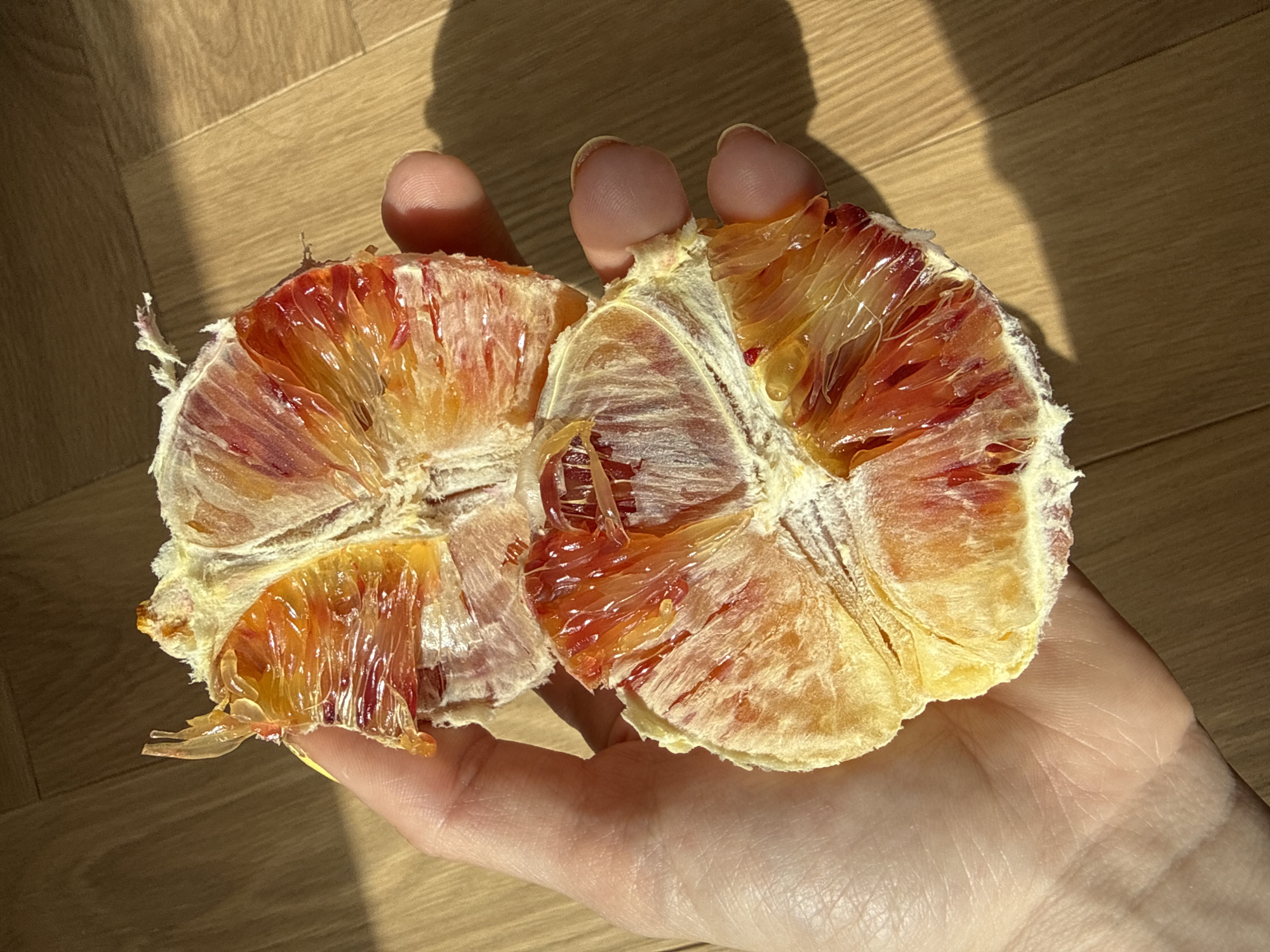A close-up shot shows a hand holding a partially peeled blood orange. The orange is split into two halves, revealing the juicy segments. The hand appears to be resting on a wooden surface. The background is blurred, but suggests a room with wooden flooring. There is no indication of the location being in Moscow, Russia.