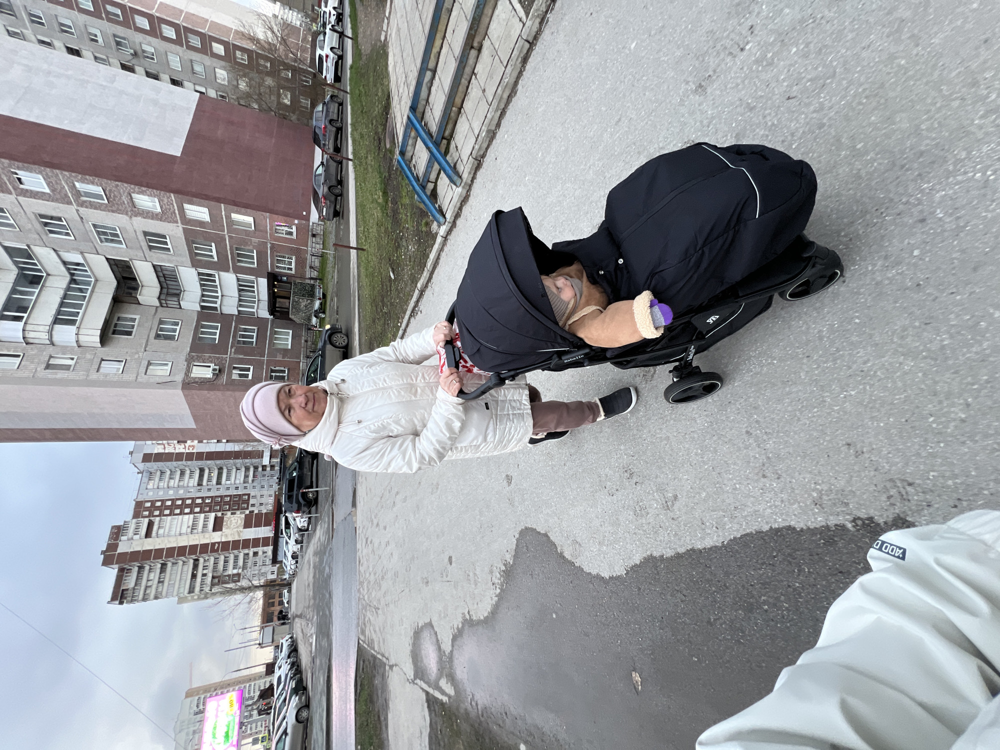 The image captures a woman pushing a baby stroller on a concrete sidewalk in an urban environment, consistent with Perm, Russia. The woman, an older adult, is seen from the waist up, wearing a white quilted jacket and a light pink knitted hat. She offers a gentle smile towards the camera. The black baby stroller, which has "Bebello" visible on its side, holds a baby or young child partially covered by blankets, with a purple pacifier in their mouth.
The setting is characterized by several multi-story apartment blocks in the background, typical of Soviet-era architecture, with grey brick, concrete, and some sections painted in brown and white. Cars are parked along the street beside the sidewalk, and sparse, bare trees suggest late autumn, winter, or early spring. The ground is damp with puddles on both the asphalt road and concrete pavement, indicating recent rain or snowmelt. The sky is overcast and grey.
In the mid-ground, a blue metal railing or staircase is visible. A distant billboard with some unreadable Cyrillic text can be seen in the far background. A label on the woman's jacket displays partial text, possibly "ADD D". The scene portrays a casual outdoor stroll under a cloudy sky.