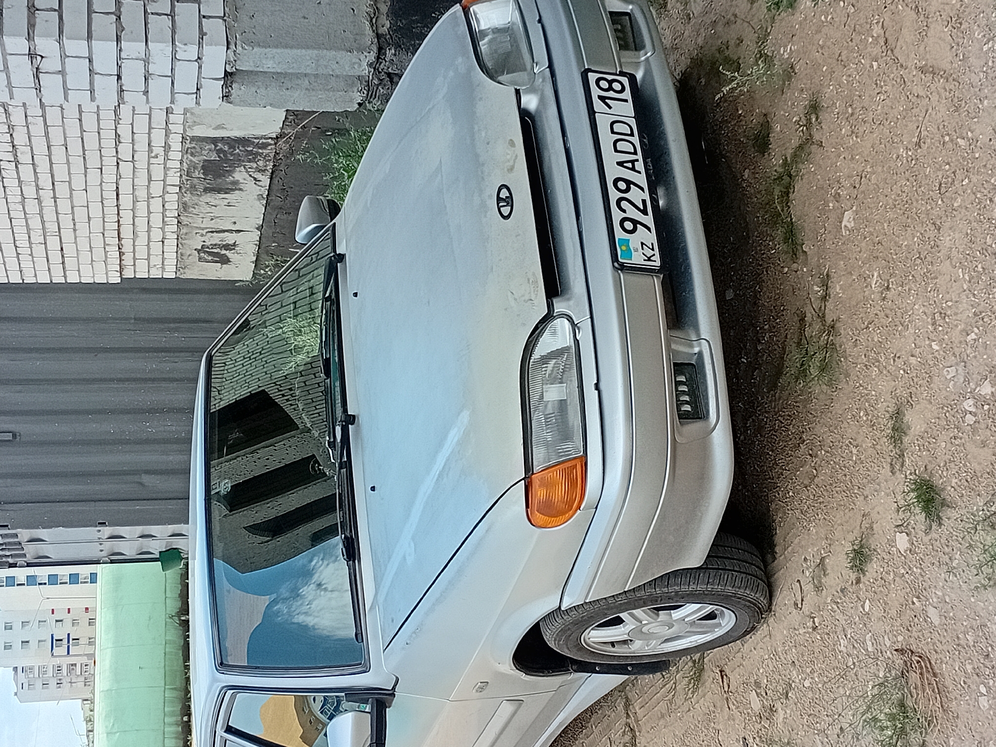 A silver sedan, a Lada 2114, is parked on a dirt and gravel lot. The car is facing forward and is slightly angled to the right. The license plate is visible and reads "KZ 929 ADD 18". In the background, there is a white brick building with a grey corrugated metal fence. Further in the distance, several tall apartment buildings are visible. The scene appears to be in the city of Semey, Kazakhstan, with the architecture and license plate indicating a Central Asian location. There are no people or animals visible in the image. The lighting suggests it is daytime.
