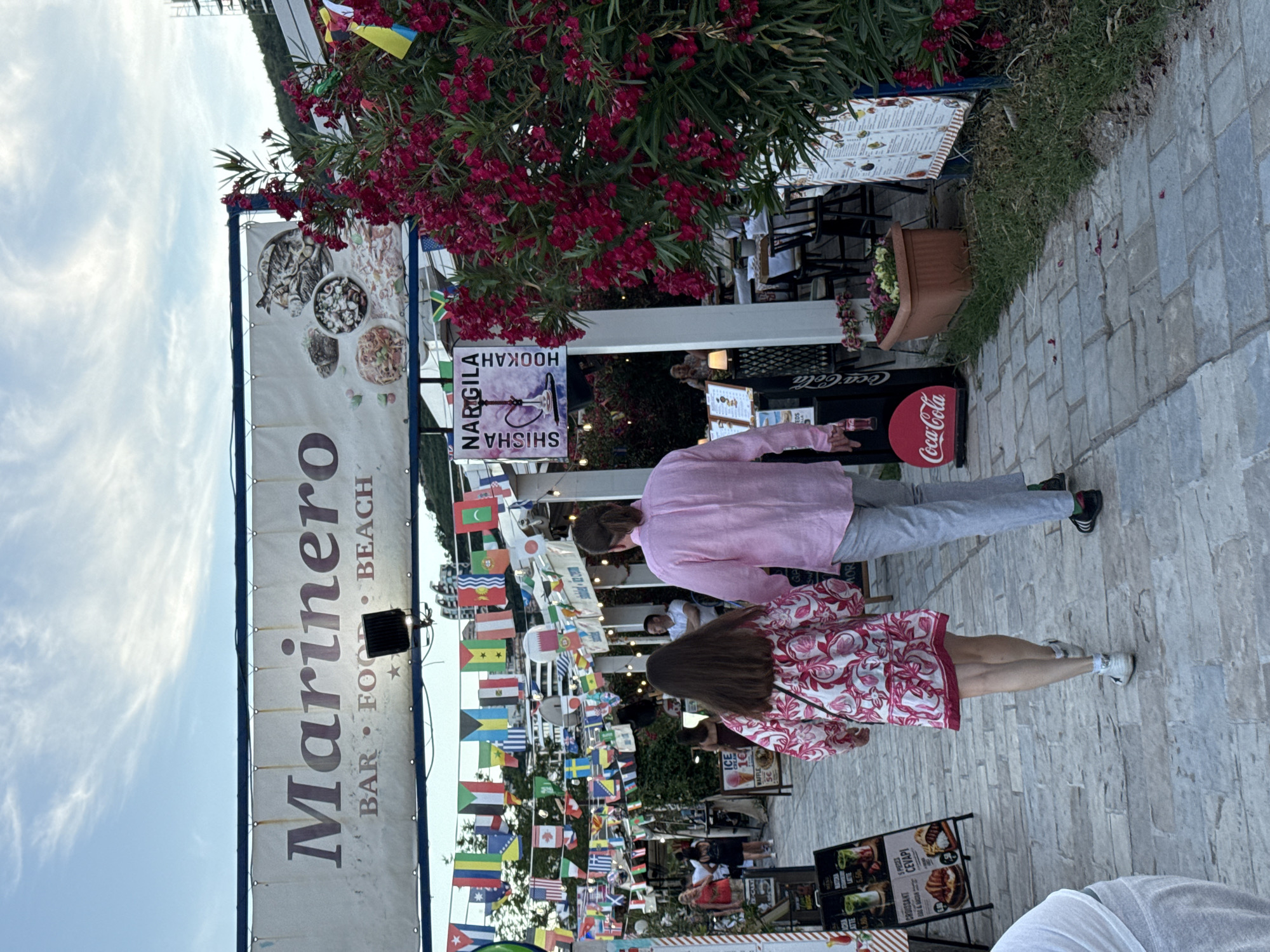 This image depicts a promenade area in Budva, Montenegro, likely in the vicinity of the Old Town or a beachfront establishment. In the foreground, a couple walks away from the camera. The woman wears a pink and white floral dress, and the man is in a pink long-sleeved shirt and grey pants. To the left, a sign advertising "Marinero BAR FOOD BEACH" is visible, with various national flags strung across the pathway above. To the right, a lush bush with vibrant pink flowers and a sign for "NARGILA SHISHA HOOKAH" are prominent. Further in the background, outdoor seating areas with tables and chairs suggest a dining or lounge establishment. Several other people are visible in the mid-ground, some walking and others seated. The pathway is paved with stone. The sky is partly cloudy.