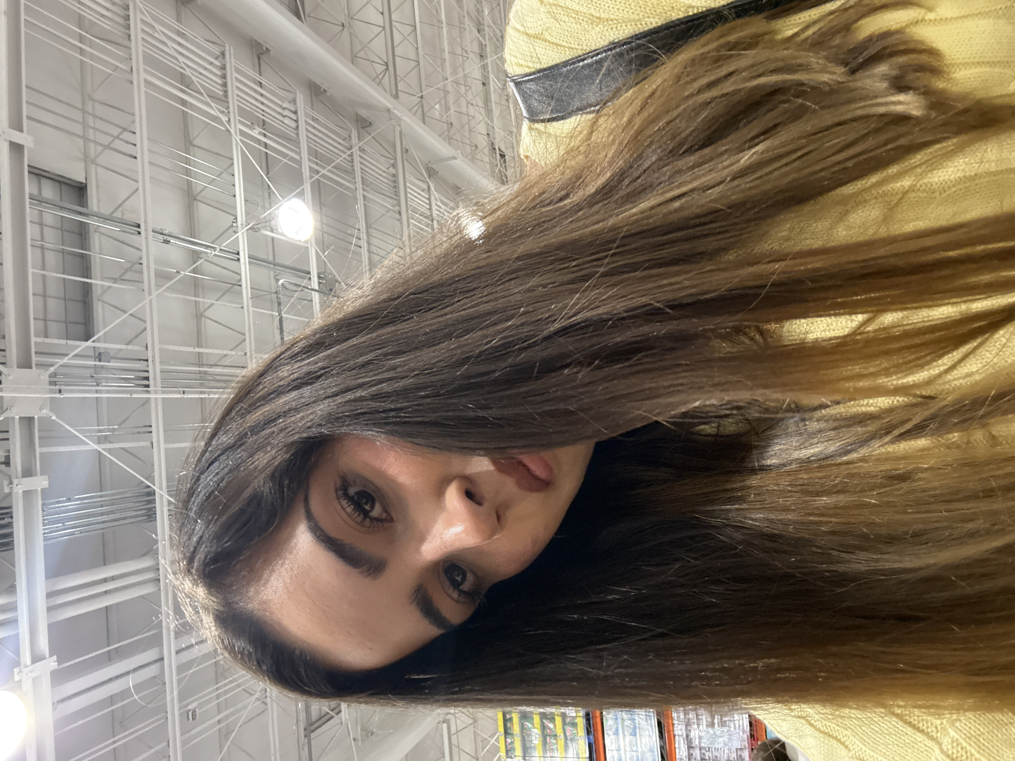 A tilted medium shot shows a woman with long, brown hair and fair skin. She wears a yellow sweater with a black strap over her shoulder. The background is an industrial-style ceiling with white metal beams and light fixtures, suggesting a warehouse or retail space. Products are visible on shelves in the distance, indicating a retail store. The location is unspecified in the city of Puebla, Mexico. No specific actions or interactions are discernible.