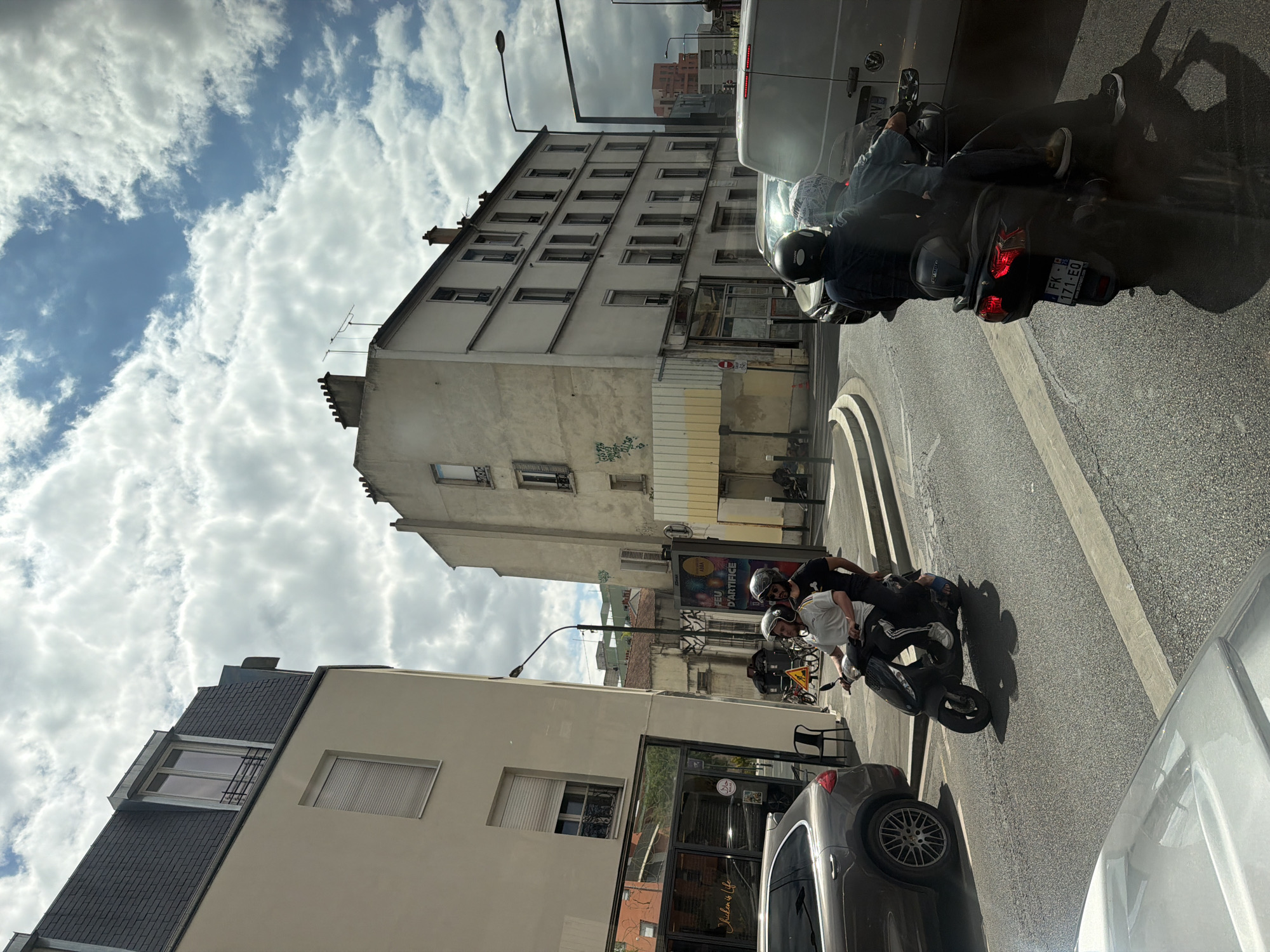 The image depicts a street scene in Alfortville, France, on a cloudy day. The main subjects are three people on a scooter, two of whom are in the foreground. The person in the front is wearing a dark helmet and a dark jacket, and the person behind them is wearing a white shirt and a helmet. A third person, mostly obscured, is behind them. They appear to be riding or stopped on the scooter.
The setting is an urban environment with buildings lining the street. On the left, there's a light-colored building with multiple windows and a modern, dark-tiled roof on an upper section. Further back, a taller, older building with a grid of windows dominates the background. The street itself is paved asphalt, with a concrete curb and a painted line indicating a lane.
Notable details include the scooter's license plate, which is partially visible and appears to read "FK 171-EQ". Shadows are cast on the ground, suggesting the sun is present, albeit partially obscured by clouds. The overall mood seems casual, possibly depicting friends or individuals commuting.
There are also glimpses of other vehicles, including a silver car in the foreground on the right and a white van further up the street. Advertisements are visible on a billboard in the middle ground. The weather appears to be mixed, with patches of blue sky visible through the clouds. The time of day is likely daytime, given the lighting conditions.