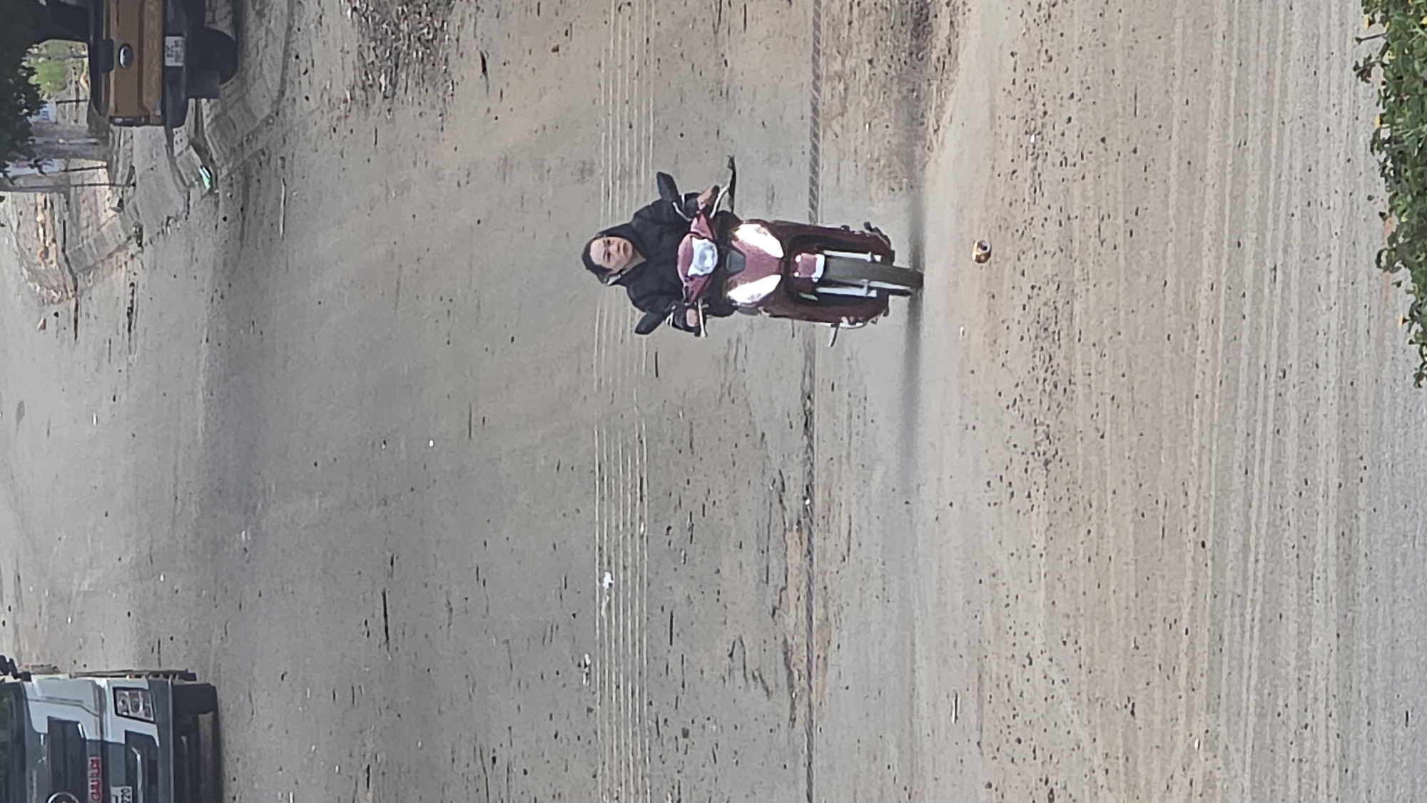 The scene is Bờ Y, Vietnam. The image presents an overhead view of a person riding a red moped-style motorbike on a road. The person is wearing dark clothing. The road appears to be concrete or asphalt with visible debris and markings. A truck is partially visible in the lower left corner of the frame. Another truck is visible in the upper left corner of the frame. Vegetation is visible on the right side of the frame.