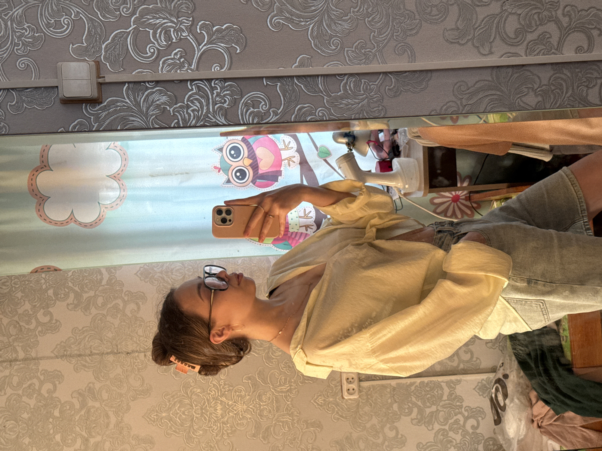 A young woman with brown hair pulled into a bun, secured with an orange clip, and wearing large black-rimmed glasses, is taking a mirror selfie in an indoor setting. She is dressed in a loose, light yellow, textured button-down shirt with rolled-up sleeves and light blue denim shorts. She is also wearing a thin gold necklace and hoop earrings. Her left hand is holding a rose-gold smartphone, angled to capture her reflection. The background of the mirror shows a light blue curtain with a pastel cloud pattern and an illustration of a pink and purple owl. To the right of the curtain, a grey wall with a white floral damask pattern is visible, with a light-colored electrical outlet. The woman's right hand is in her pocket. The environment appears to be a bedroom, with a wooden dresser or table visible in the reflection behind her, displaying a white desk lamp and some items on its surface. Clothing is also piled on a wooden surface to the left of the dresser. The overall impression is of a casual, personal moment captured in a domestic space. There is no indication this scene is located in the city of Kemul, Russia; it appears to be a standard bedroom setting.