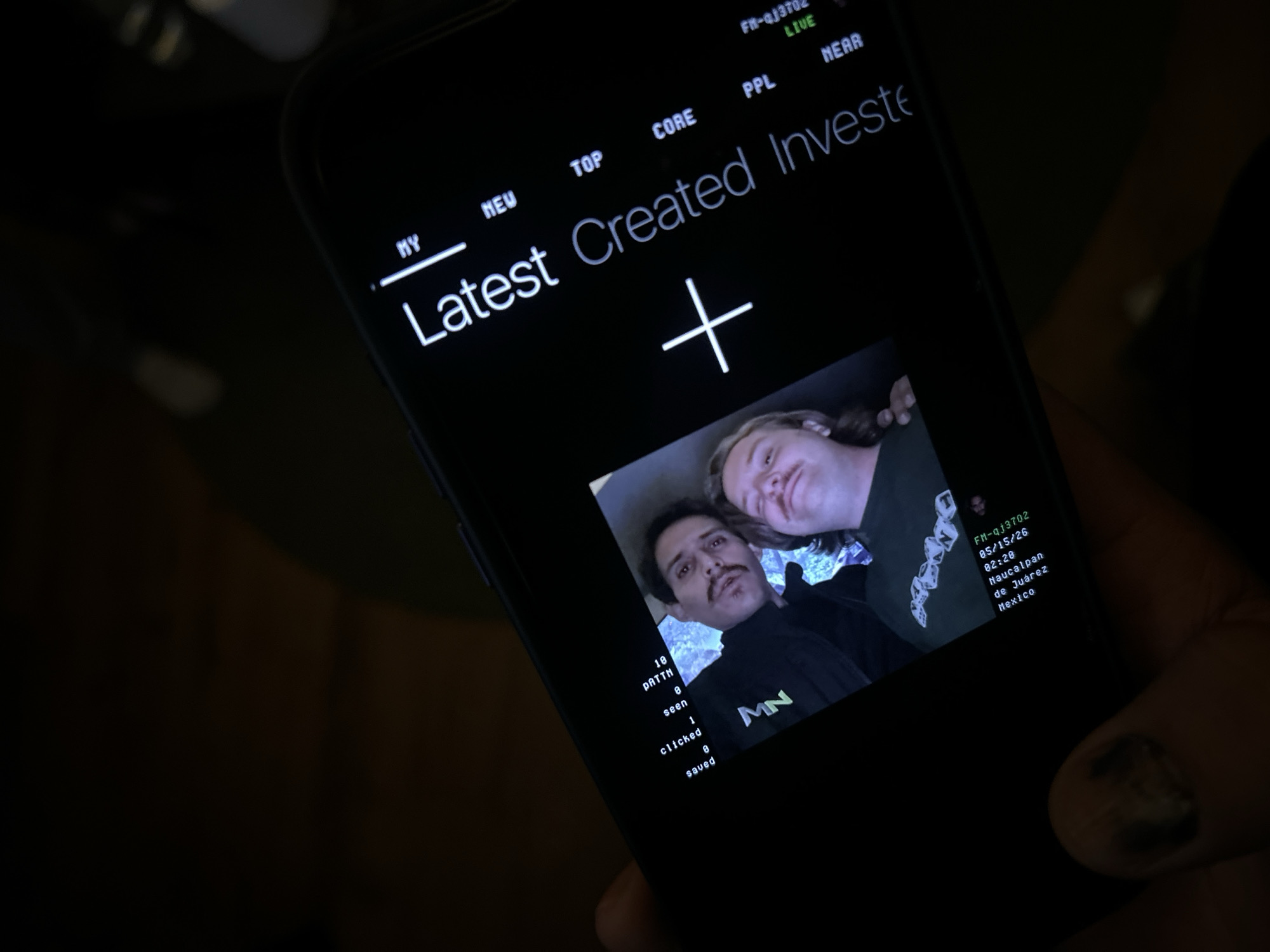 A mobile phone screen displays a social media-like application showing a selfie of two individuals. One person has a mustache and is wearing a black shirt with a green and black logo "MN." The second person is seen in the background, slightly out of focus. The application interface shows the labels "Latest Created Investe" and a "+" icon, indicating a content creation or adding feature. Text on the screen includes "PATTN 10", "seen 1", "clicked," and "saved 0." Location information displays "Naucalpan de Juárez, Mexico," and the alphanumeric code "FM-qj3702 05/15/26 02:20." The screen also has labels such as "MY," "NEU," "TOP," "CORE," "PPL," and "NEAR." The phone is being held in someone's hand. Huixquilucan de Degollado is not mentioned on the screen; only Naucalpan de Juárez, Mexico, is visible.
