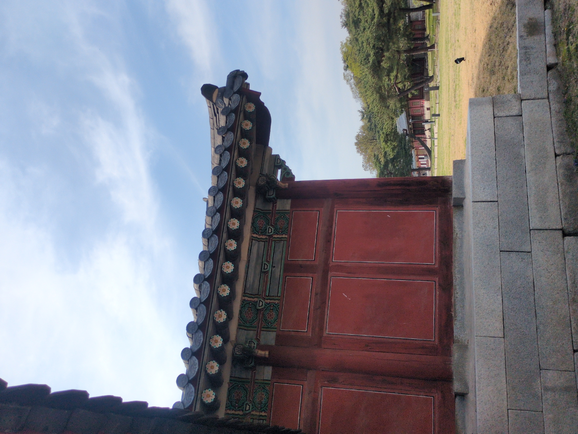 A low-angle shot captures a fragment of traditional Korean architecture. The structure features a red painted wooden wall, adorned with rectangular paneling and decorative detailing. Above the wall is a traditional roofline with dark gray tiles arranged in a floral pattern.
To the right of the building is a gray stone wall, built from stacked rectangular blocks.
In the background, a grassy expanse is visible, dotted with scattered trees and small buildings with red-painted details. A dark bird is on the grass.
The scene suggests a historical site in Seoul, South Korea.