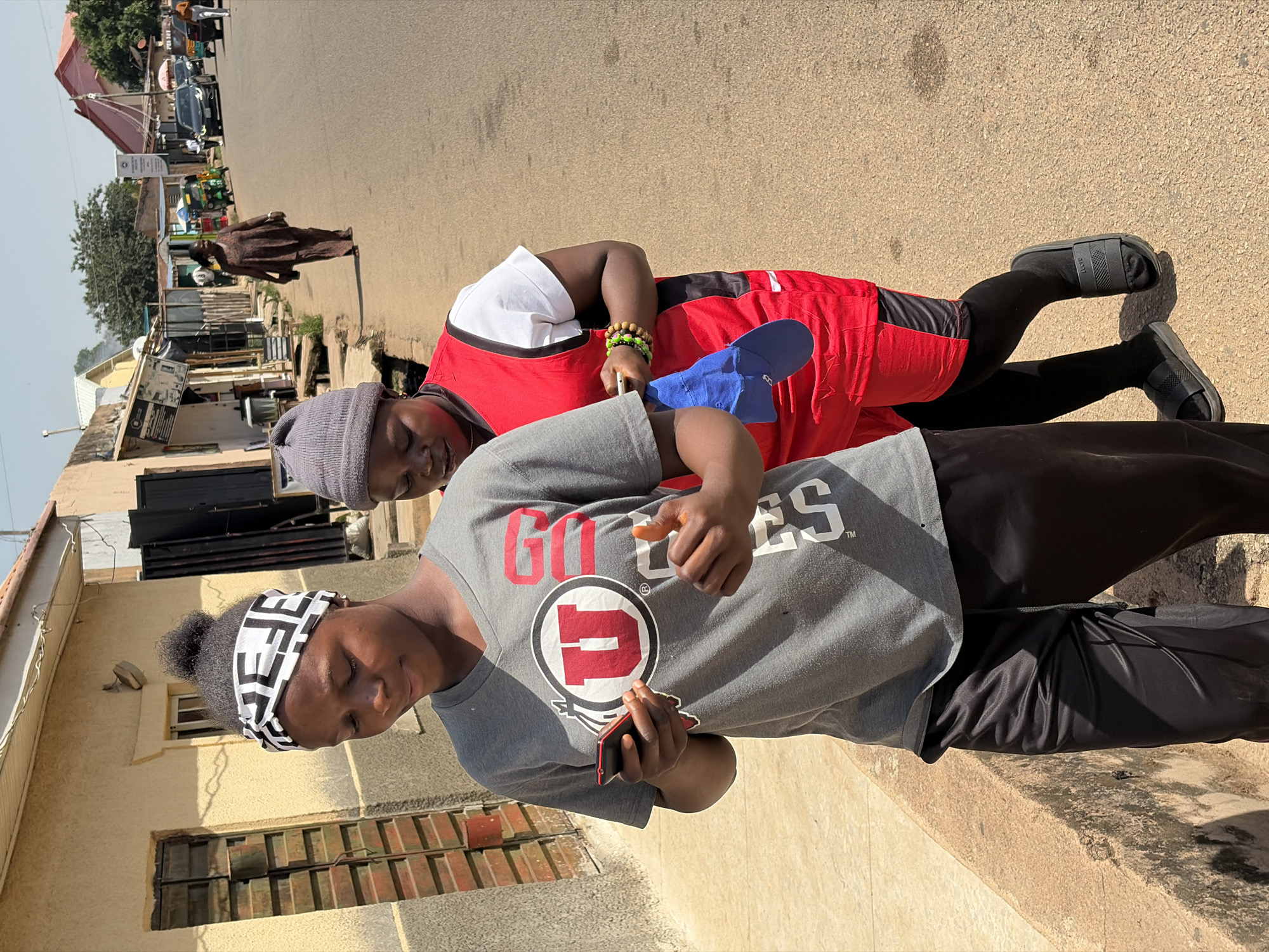 Two women are standing close to each other in an urban environment, possibly in Jos, Nigeria. One woman wears a gray T-shirt with a "U" logo, black pants, and a patterned headband, holding a phone and pointing with her other hand. The other woman wears a gray beanie, a red apron over a white shirt, black leggings, and sandals, holding a blue object. They are positioned next to a building and a street with passing cars and pedestrians. The environment includes buildings, shops, and general urban infrastructure.