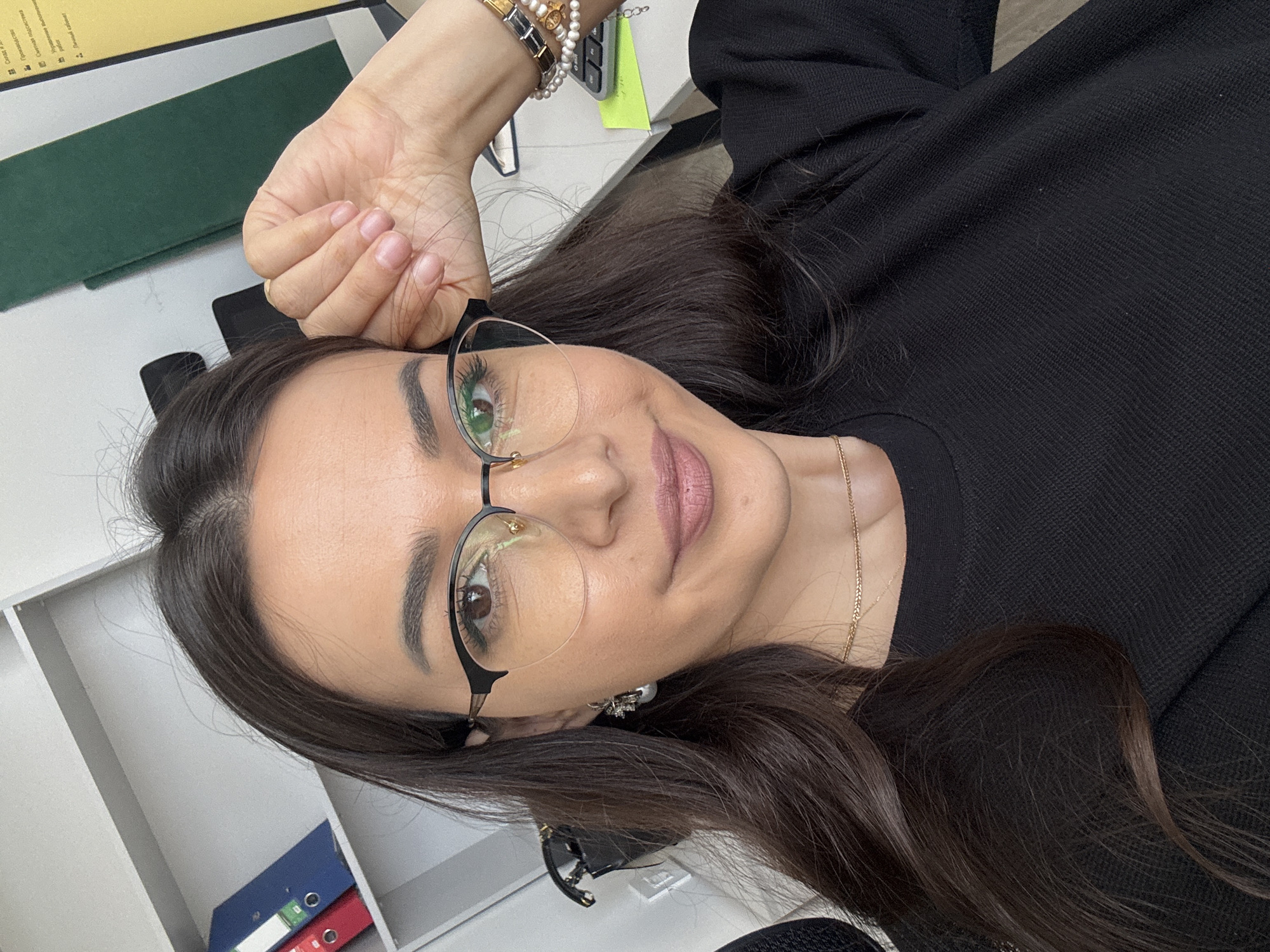 A woman with dark hair and glasses is taking a selfie in an office environment. She is wearing a black long-sleeved shirt and a gold necklace. She has a pearl earring and several bracelets on her left wrist. Her right hand is resting on her forehead. In the background, there are shelves with binders, a green divider, a computer monitor, and a keyboard.