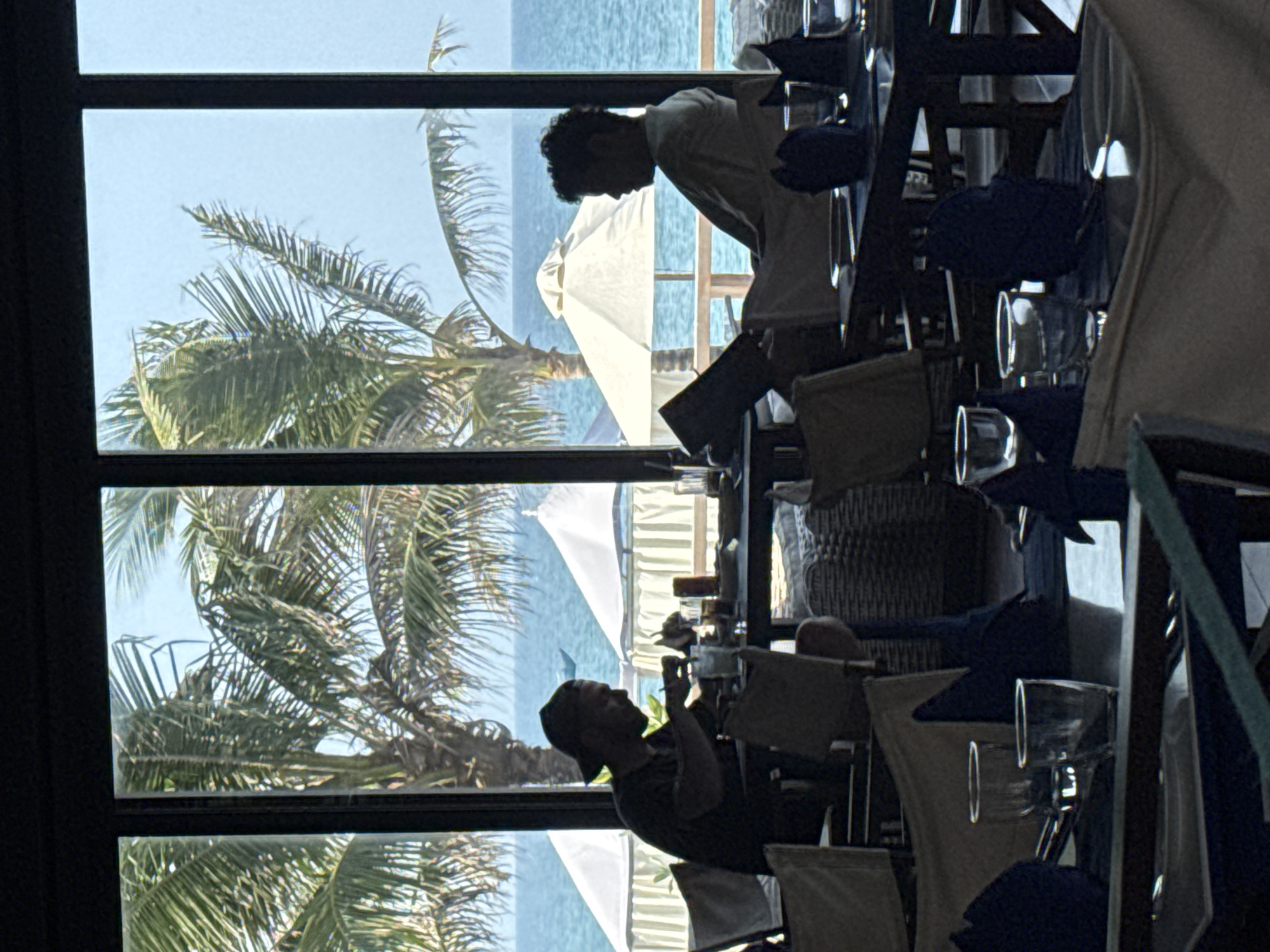 The image shows an interior scene of a restaurant or dining area with a large window offering a view of a tropical beach. Through the window, palm trees, the ocean, and beach umbrellas are visible, suggesting a coastal location. Inside, there are people at tables. One person in a light-colored shirt appears to be a server or waiter, perhaps interacting with customers. Another person at a table seems to be using a cell phone. The tables are set with plates, napkins, glasses, and blue placemats. The image suggests a relaxed, seaside dining atmosphere. No specific evidence indicates it's Đà Nẵng, Vietnam, but the presence of tropical elements in the image makes a location in Vietnam or another similar tropical seaside setting probable.