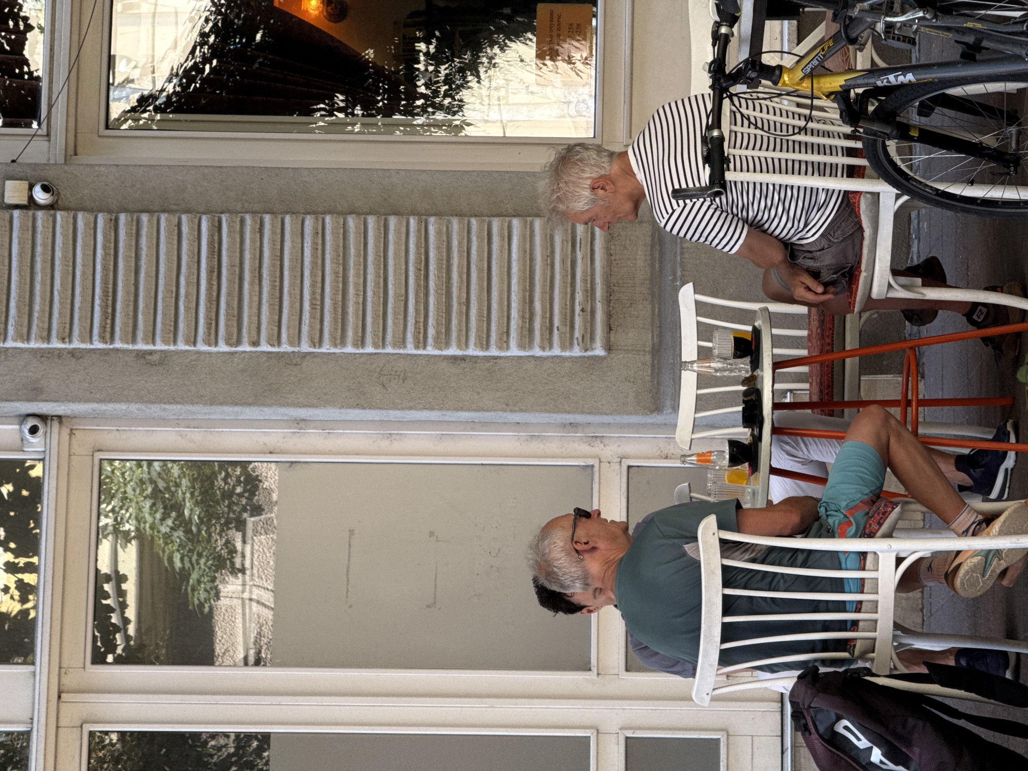 Two men and part of a third man are seated at tables outside a building with large glass windows. The man on the left, wearing a green t-shirt and blue and orange shorts, is facing away from the camera, seated on a white chair. He has a bag with "HEAD" written on it to his left. In front of him is a small table with drinks. The man in the middle, wearing a grey t-shirt, is mostly obscured by the man in green. The man on the right, wearing a striped t-shirt, is facing left, looking away from the camera. He is seated on a white chair. A bicycle is parked to his right. The building appears to be a cafe or restaurant. The scene is located on a street in the city of Belgrade, Serbia, likely in a commercial or pedestrian area given the outdoor seating and the presence of a bicycle.