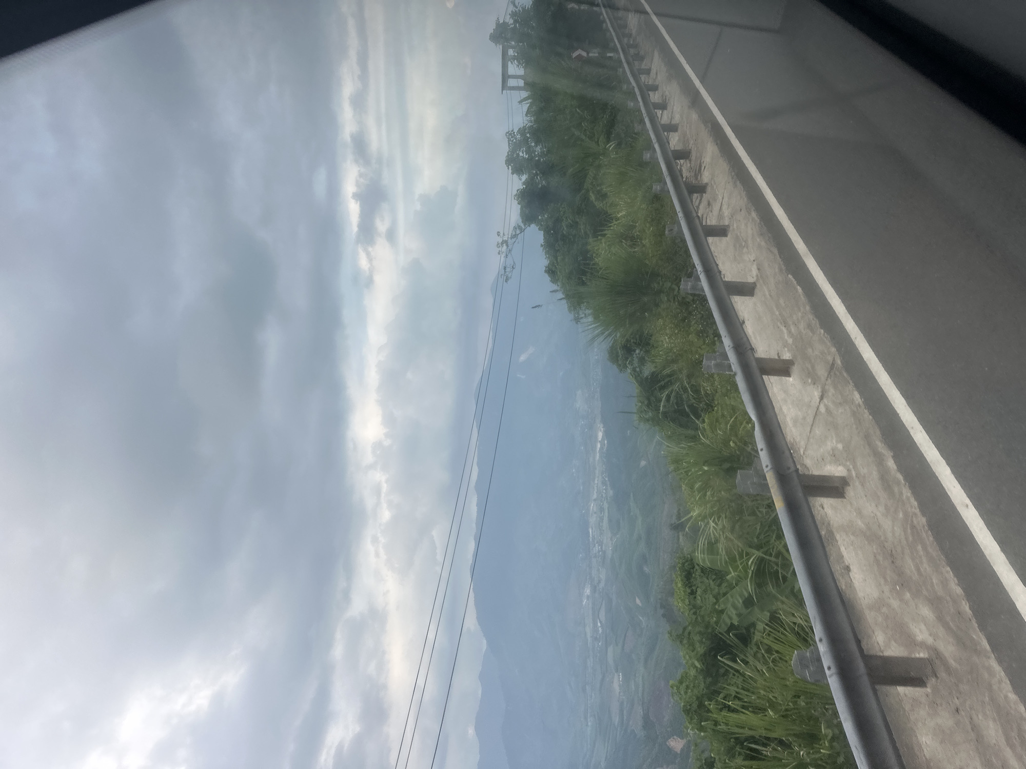 The image shows a view from a roadside on a cloudy day, possibly in Tay Khanh Vinh, Vietnam. A grey sky filled with striated clouds is visible. Below the sky is a mountain range with vegetation and a town or settlement nestled within the valley. A concrete barrier lines the road. Power lines stretch across the landscape.