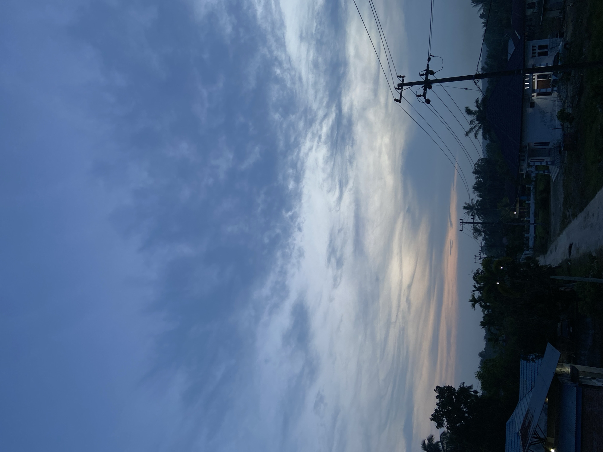 The image depicts a twilight scene in Candirejo, Indonesia. The sky is a blend of deep blues and lighter hues near the horizon, with scattered clouds. In the foreground, a dirt road leads into a village. To the right, a white house with a dark roof is illuminated by interior lights, and a utility pole with power lines stands nearby. On the left, the roof of a building is visible, and silhouetted trees dot the landscape. The overall atmosphere is quiet and serene, characteristic of early evening in a rural Indonesian setting.