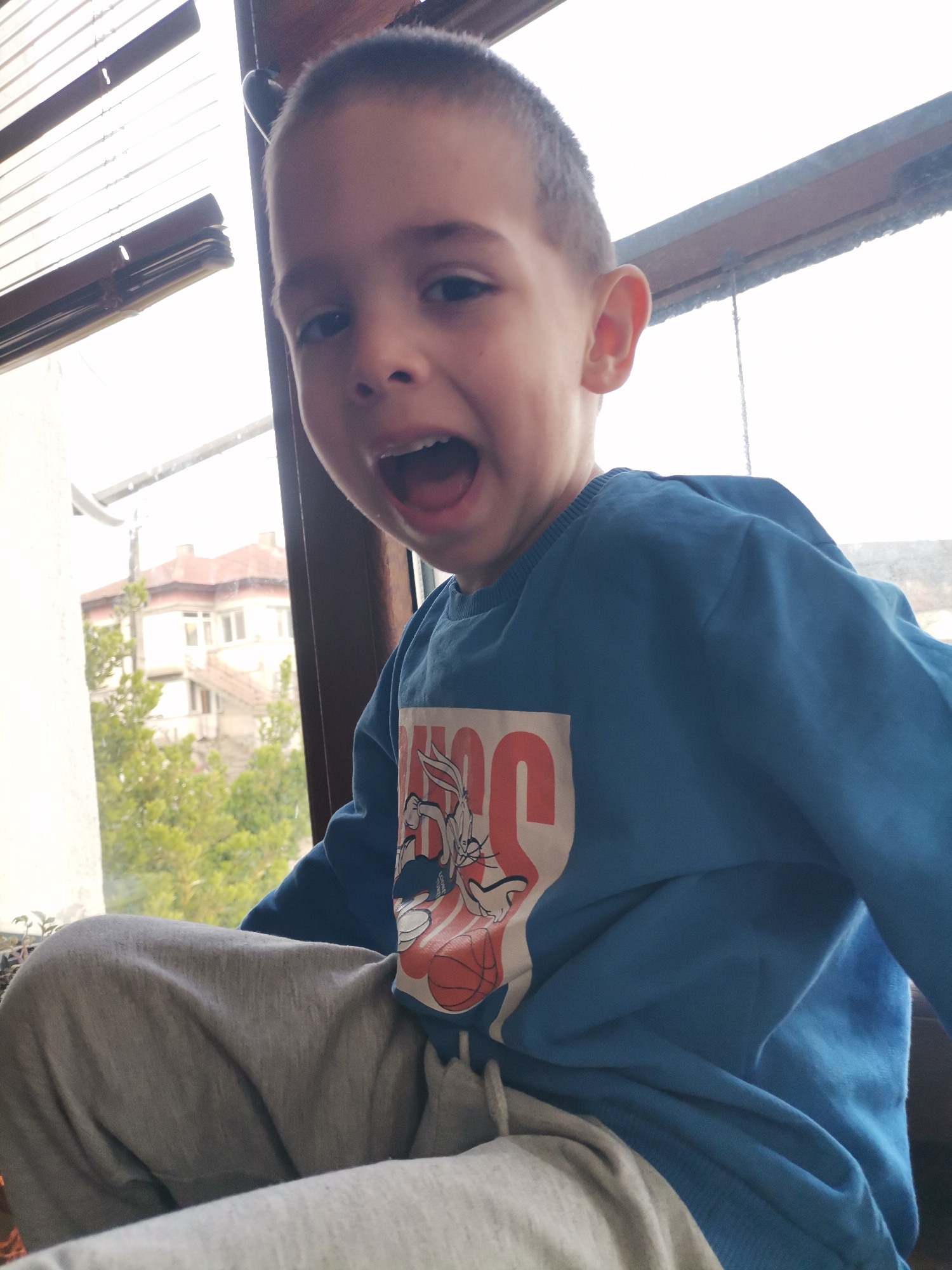 A young boy with short hair is sitting indoors, possibly on a window sill, with a building and greenery visible outside the window. He is wearing a blue long-sleeved shirt featuring a Bugs Bunny design and gray sweatpants. His mouth is open, and he appears to be calling out or making a loud expression. The location is in the city of Unknown, Unknown.