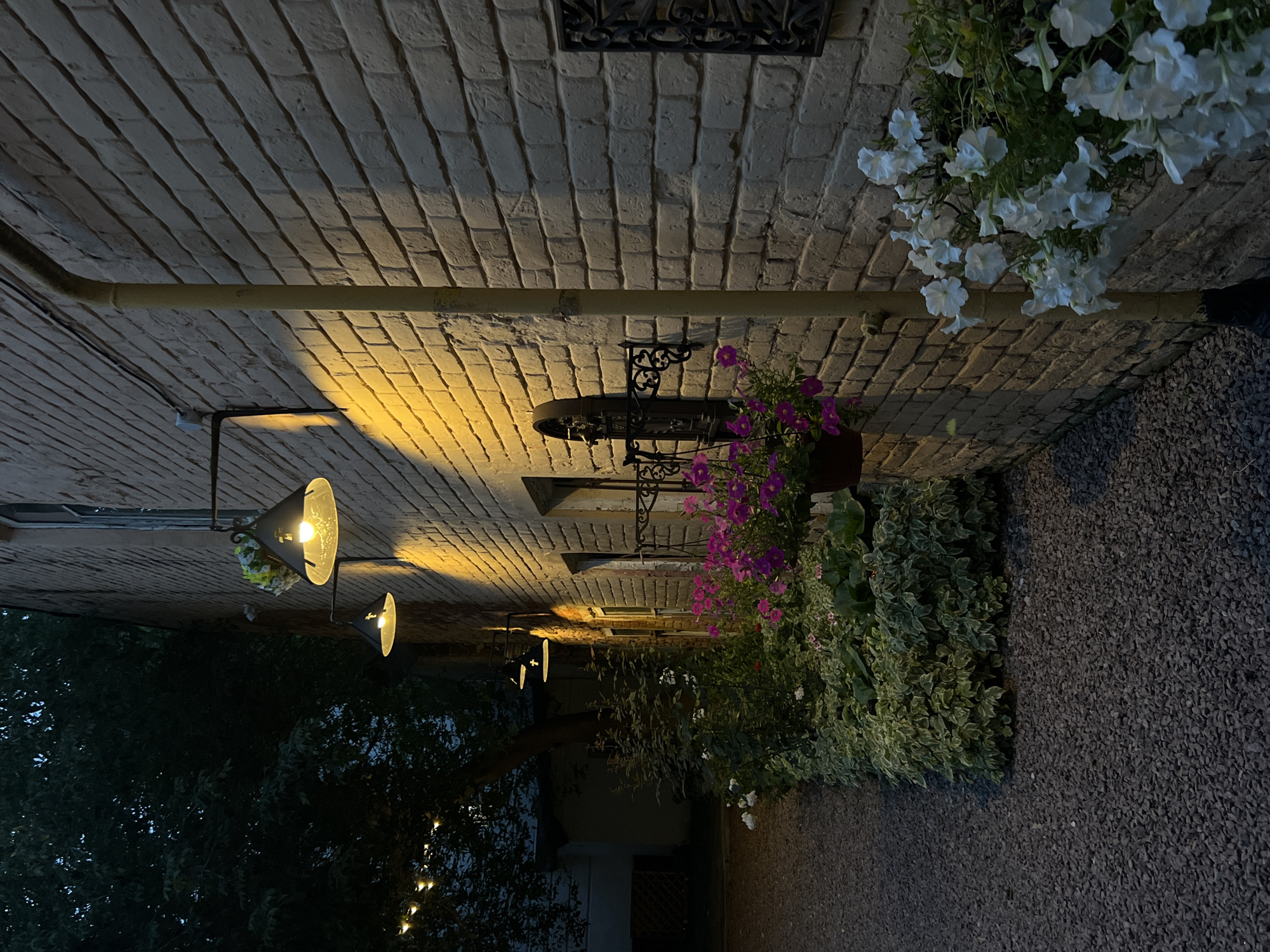 The image shows a brick wall with exterior lights, potted flowers, and gravel. The brick wall is light yellow and appears aged. Several dark-colored lamps with wide shades are mounted on the wall, emitting a warm yellow light. In front of the wall, a row of lush green plants with some white and pink flowers in pots are visible. The ground is covered with dark grey gravel. The scene suggests an outdoor area, possibly a courtyard or garden, at dusk or in the evening, with artificial lighting illuminating the wall and plants. The overall impression is one of a rustic or charming outdoor setting. There are no people or visible actions occurring in the image.