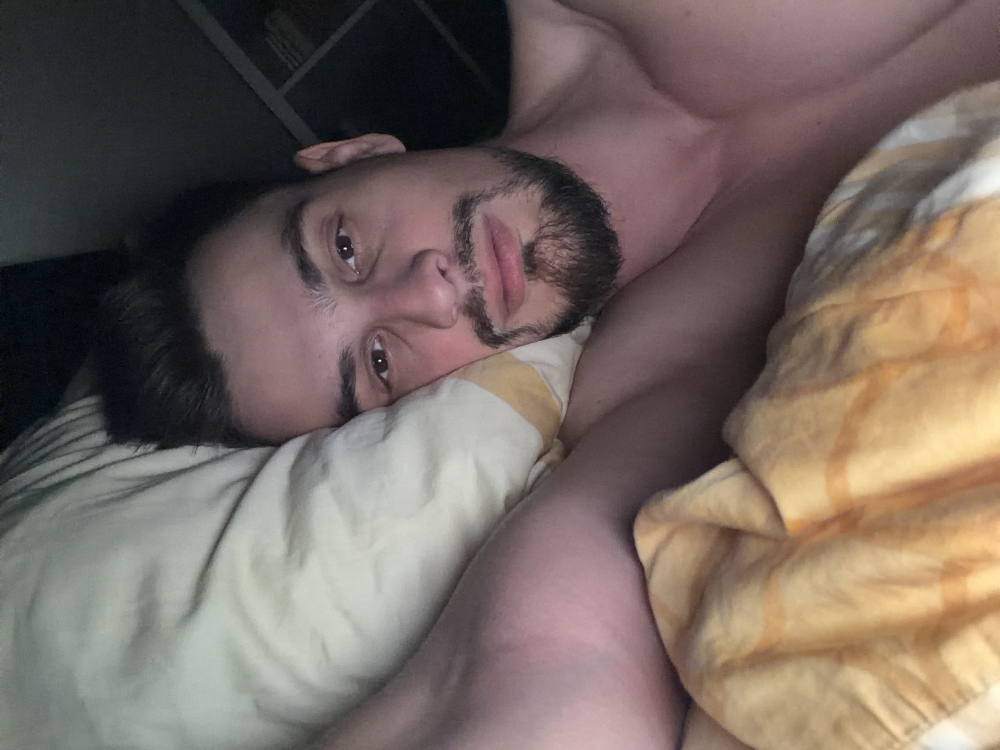 A man with dark hair and a beard lies on his side in a bed, looking at the camera. He is resting his head on a pillow with a yellow and white striped pattern. He is partially covered by a yellow blanket with a similar striped pattern. The background is dark and out of focus, but appears to be a room with shelves or furniture on the right side. The image was taken in Krasnoyarsk, Russia.