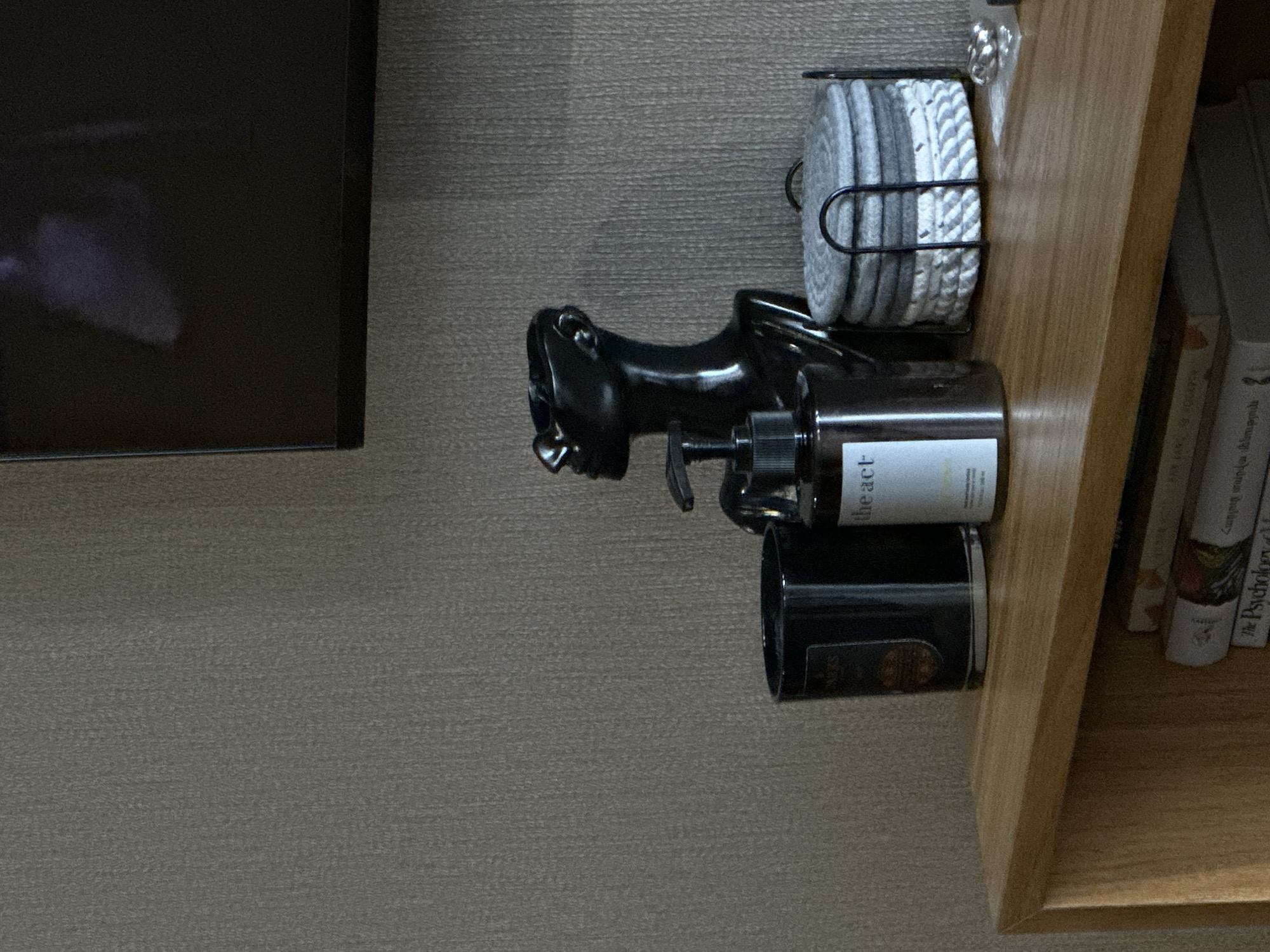 The image depicts a portion of a room's interior. A flat-screen television is partially visible on the left. Next to it, a light brown wooden shelf contains a stack of gray and white circular cloths in a metal holder, a black decorative sculpture resembling a human head, a bottle labeled "the act", a black canister, and a row of books. The location in the city of Yerevan, Armenia cannot be determined from this image.