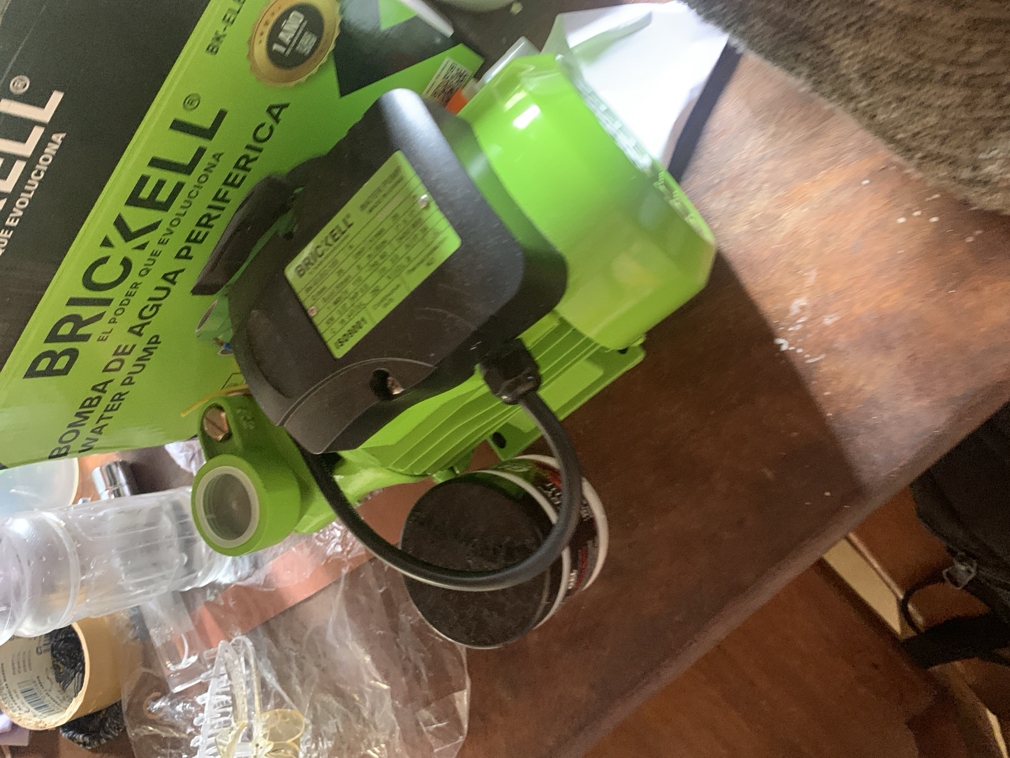 The image depicts a cluttered wooden table surface with various objects. A bright green and black water pump is prominently featured, with a green housing and a black motor unit. Next to it, there are two small, dark-colored containers, one with a black lid and the other partially visible. A clear plastic bottle filled with liquid is also present, along with a roll of tape and some clear plastic sheeting. In the background, a portion of a green box with text related to "BRICKELL WATER PUMP" is visible, along with a gold circular sticker indicating "1 AÑO". The table surface itself appears worn and has some scattered white debris. The overall setting suggests an indoor environment, possibly a workshop or storage area. There is no indication of the location in Santa Ana, Venezuela, or any people present in the image.