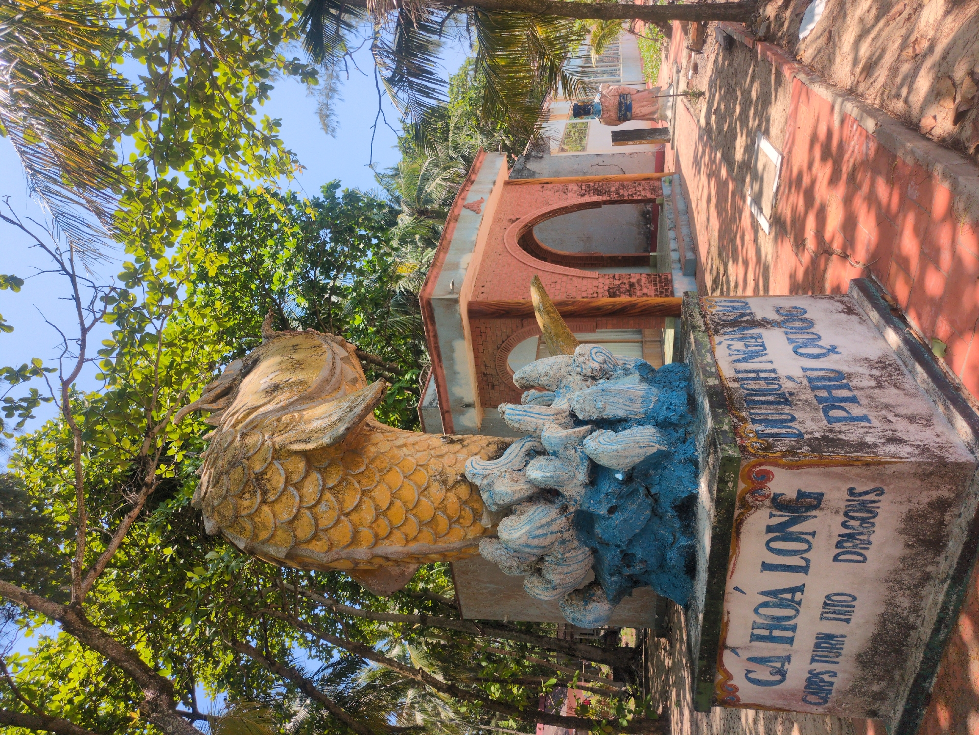 The image shows a statue of a carp transforming into a dragon (Cá Hóa Long) in Phú Quốc, Vietnam. The statue is mostly yellow with blue and white waves surrounding it. The statue sits atop a concrete base with the name "Cá Hóa Long" written in Vietnamese and English. Behind the statue is a red brick building with arched openings. A figure stands near the building. The environment includes trees, blue sky, and a path made of red tiles.