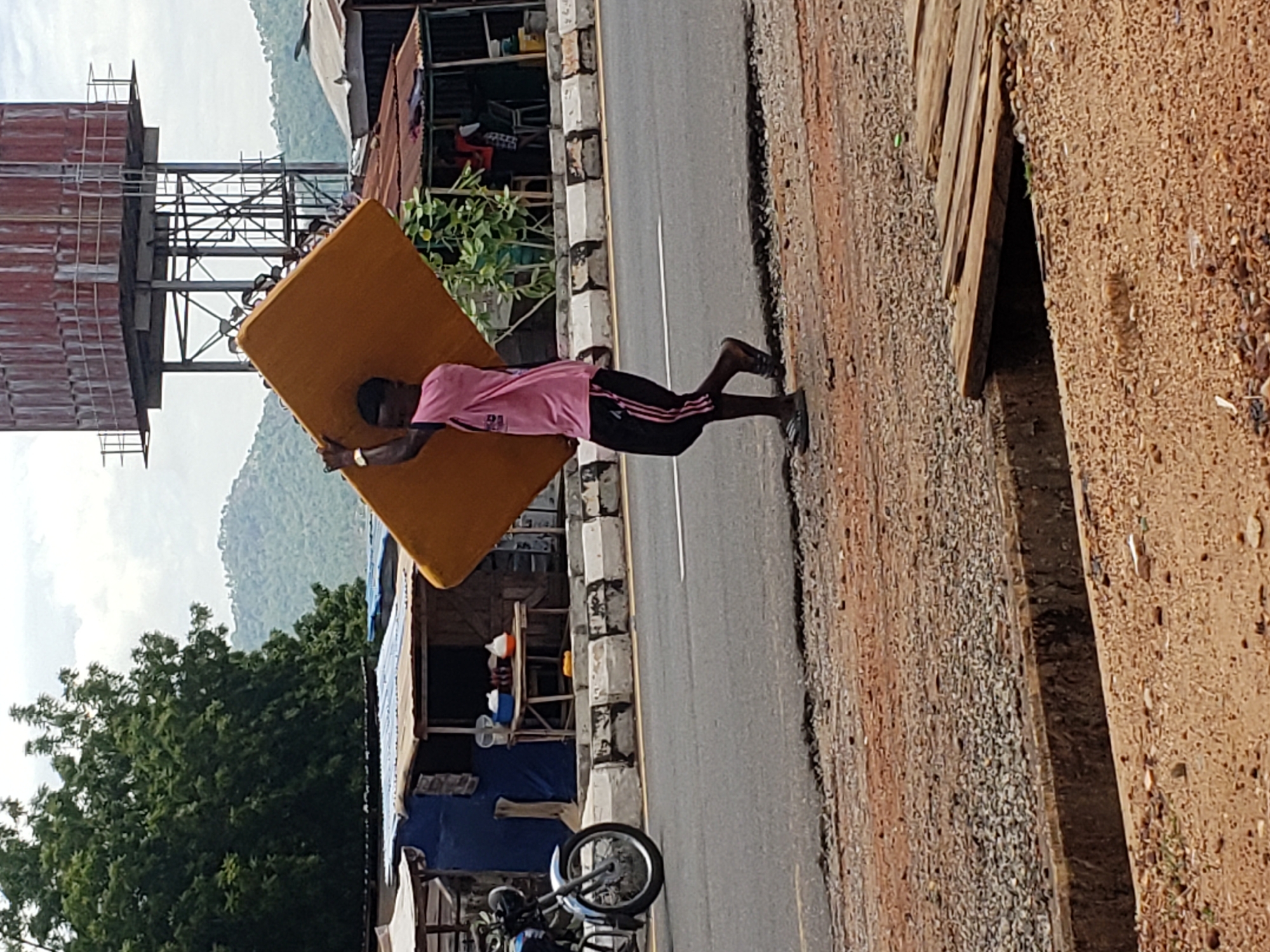 The image depicts an outdoor scene along a paved road, likely in Adankolo, Nigeria, featuring a person transporting a mattress during daytime. A dark-skinned individual, appearing male, is the central subject, walking from left to right along the road's shoulder. They are carrying a large, rectangular, golden-yellow mattress horizontally, held against their body with outstretched arms, indicating physical effort. The person wears a pink t-shirt with faint white numbers, possibly "23" or "22," on the front, black athletic shorts with three white stripes down the side, and dark footwear.
To the right of the person is a dirt and gravel shoulder. To the left, a grey paved road is visible, marked with a single white line. The road is bordered by a curb made of alternating black and white stone blocks. Further left, a motorcycle is partially parked near a makeshift roadside stall, constructed from wooden frames, corrugated metal, and a blue tarp. Various containers and items are visible on shelves inside the stall.
In the mid-ground on the far left, a prominent reddish-brown corrugated metal water tank stands on a complex metal scaffolding structure. The background features lush green trees and undulating mountains or hills under a bright, possibly overcast, sky, suggesting clear weather conditions during the day. The scene captures a moment of daily activity involving the transport of a bulky item in a semi-urban or rural setting.
