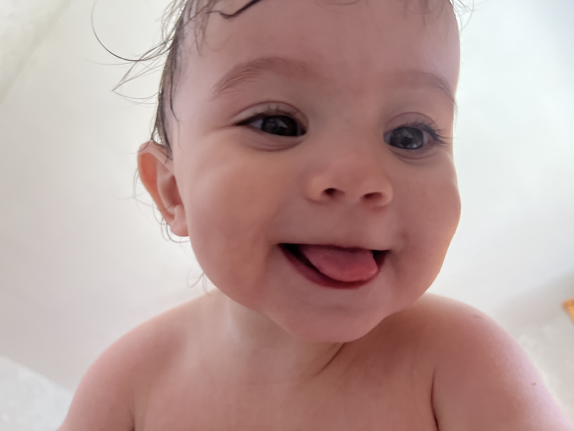 A joyful close-up portrait of a baby, likely an infant or young toddler, taken in Elena, Bulgaria. The baby has fair skin and dark, wet hair, which is styled in wispy curls, suggesting a recent bath or water play. The low-angle shot captures the baby's face and bare shoulders, filling most of the frame. They are looking directly at the camera with large, dark, expressive eyes, and a wide, happy smile. Their tongue is playfully sticking out, adding to the mischievous and cheerful expression. The background is uniformly bright and light-colored, indicating an indoor setting, possibly a bathroom, with plain walls. No text is visible in the image.