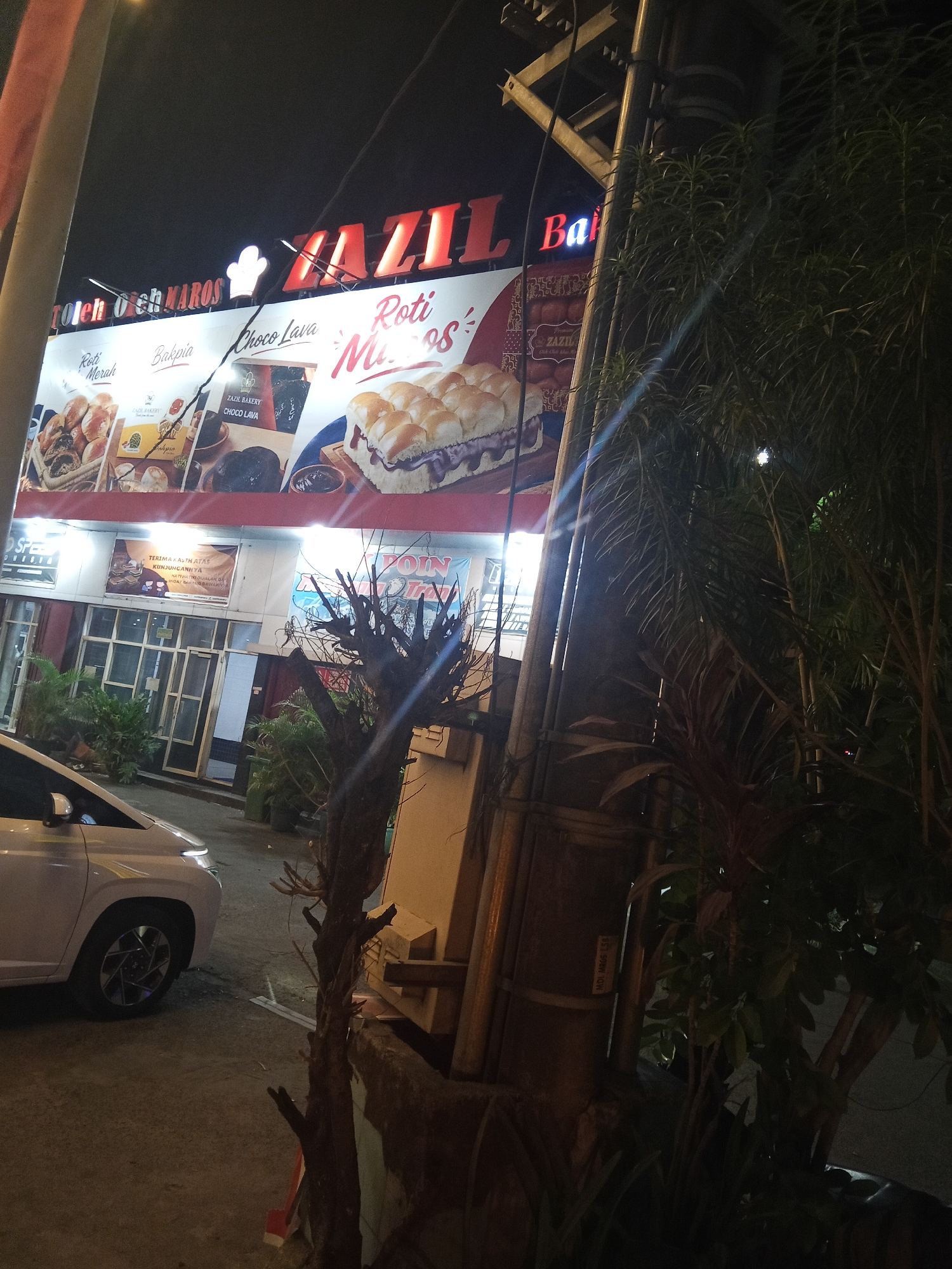 The image depicts a street scene at night in Hasanuddin, Indonesia, likely in front of a bakery or food establishment. Dominating the upper portion of the image is a brightly lit sign that reads "ZAZIL Bakery". Beneath this main sign, a larger advertisement showcases various baked goods. There are pictures of Roti Merah (red bread), Bakpia, and Choco Lava, as well as a close-up of a sandwich filled with what appears to be chocolate.
In the foreground, a white car is parked on the side of the road, its headlights illuminated. The street is paved, and there are some plants and trees visible, including a prominent, bare-branched tree to the right of the frame, partially obscuring the building. The building itself has large glass windows and doors, and some of the interior lights are on. A banner is visible hanging over the entrance that reads "TERIMA KASIH ATAS KUNJUNGANNYA" which translates to "THANK YOU FOR YOUR VISIT". Another banner with blue lettering is partially visible on the right side of the building. The overall impression is a commercial area at night, with the bakery being the focal point.