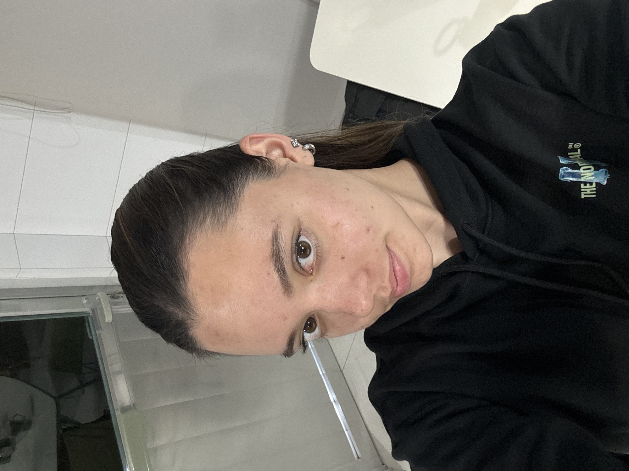 A woman is depicted in an indoor setting, likely a casual space such as a home. The image is oriented sideways, showing her face and upper torso. She has dark hair pulled back and is wearing a black hoodie. Her face shows some skin imperfections. She is looking directly at the camera with a neutral to slightly smiling expression. Her right ear is visible, adorned with a silver-colored hoop earring in the cartilage and a square stud in the lobe.
In the background, a white tiled wall is visible, along with what appears to be a window or glass door with horizontal blinds or shades on the left side. A white surface, possibly a table or countertop, is partially visible on the right. The lighting is soft and even, suggesting either diffused natural light or artificial indoor lighting.
On the front of her black hoodie, a partial logo or text is visible. The words "THE NO MAL" can be discerned, possibly part of "THE NORMAL" or a similar phrase, accompanied by a stylized graphic in shades of light blue and green, possibly representing the letter "N".
The scene suggests a casual moment, perhaps a selfie taken indoors in San Bernardino Tlaxcalancingo, Mexico.