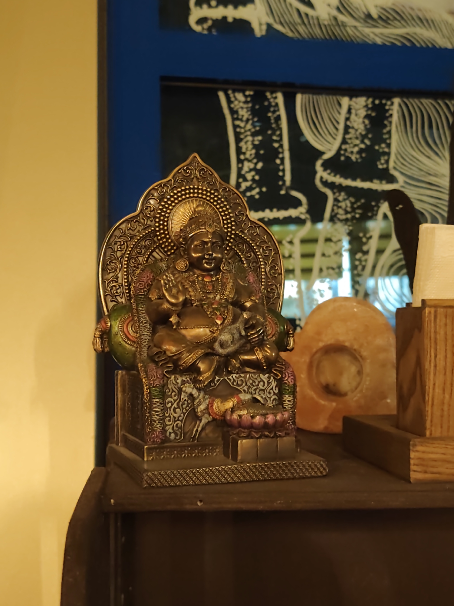 A close-up, eye-level shot shows a small, ornate statue of a seated figure, likely a deity or religious icon. The statue is made of a dark, metallic material, possibly bronze or brass, and is intricately detailed with necklaces, jewelry, and elaborate clothing. The figure is seated on a throne-like structure that is also heavily ornamented. To the right of the statue, there is a large, translucent, orange object, possibly a salt lamp, and a wooden stand holding a white object, which could be a candle or a light fixture. The background features a blue border and a dark surface with white, abstract patterns. The lighting is warm and focused on the statue, creating a sense of reverence. The image appears to be taken indoors, in a dimly lit environment. The statue is positioned on a dark wooden shelf. The overall impression is one of a shrine or a decorative display of religious or cultural artifacts. There are no people visible in the image. The scene is devoid of any specific indicators that would pinpoint its location in Sochi, Russia.