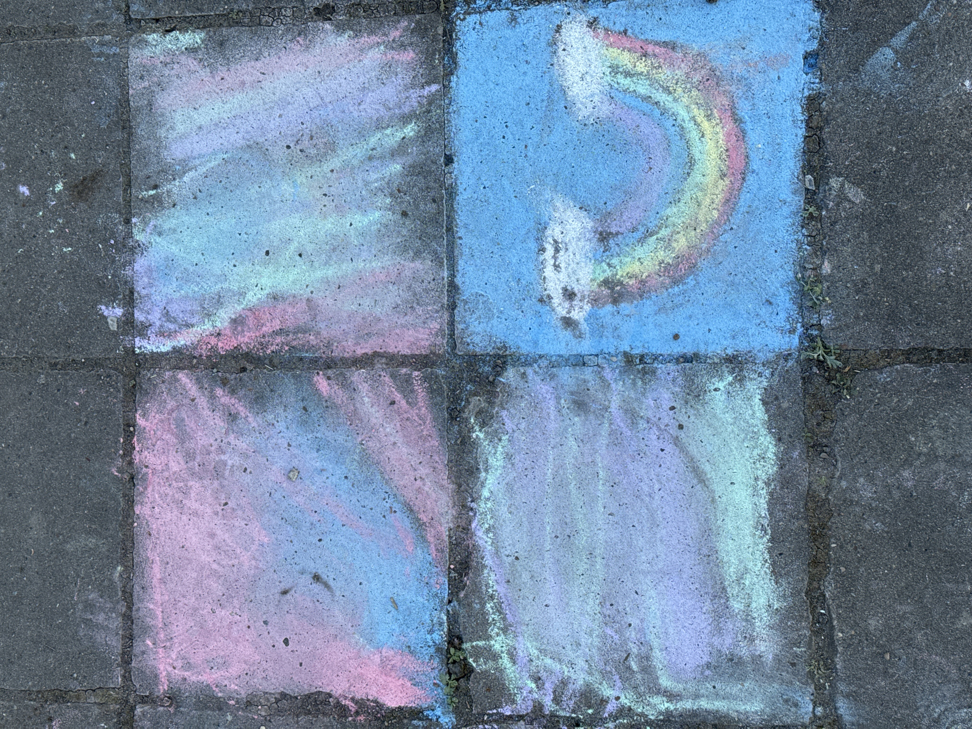 The image depicts a section of paved walkway, possibly concrete tiles, colored with chalk. The scene is divided into four tiles. The upper right tile features a rainbow drawn in chalk. The remaining tiles are colored with pastel hues of blue, pink, and lavender, creating abstract patterns. The setting is unknown and cannot be determined to be in Chaikovsky, Russia from the image. There are no people visible in the picture.