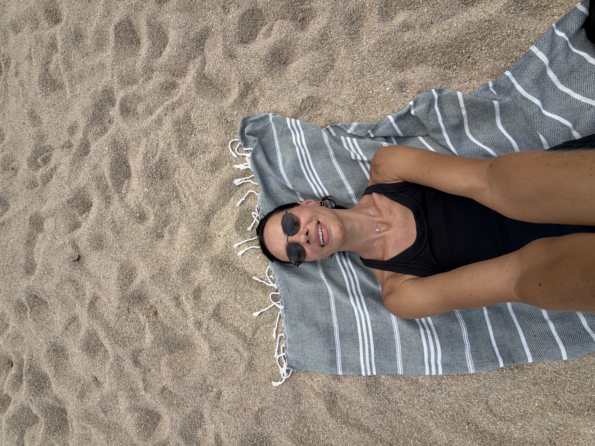A woman lies on her back on a beach towel on a sandy beach. The towel is grey and white striped with tassels. The woman is wearing a black tank top and sunglasses. Her arms are resting by her sides. The background is sand. The location is Baan Chaweng, Thailand.