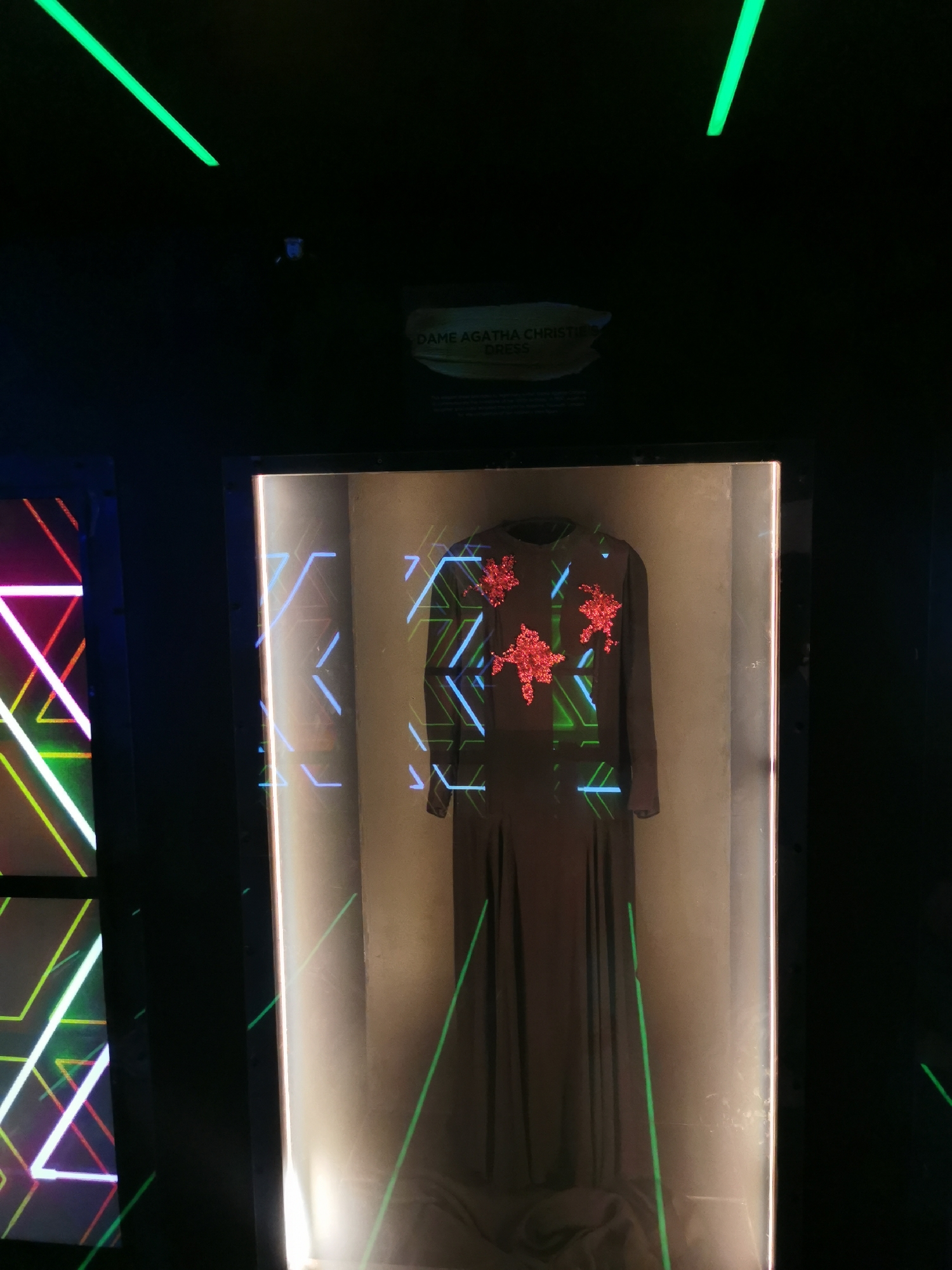 The image shows a dress displayed in a glass case. The dress is dark in color with red flower-like embellishments on the chest area. The case is illuminated with neon lights. A sign above the case reads "DAME AGATHA CHRISTIE DRESS". Another case with neon lights is to the left. The environment is dimly lit and appears to be a museum exhibit. The location is implied to be in London, United Kingdom, due to the reference to Dame Agatha Christie.