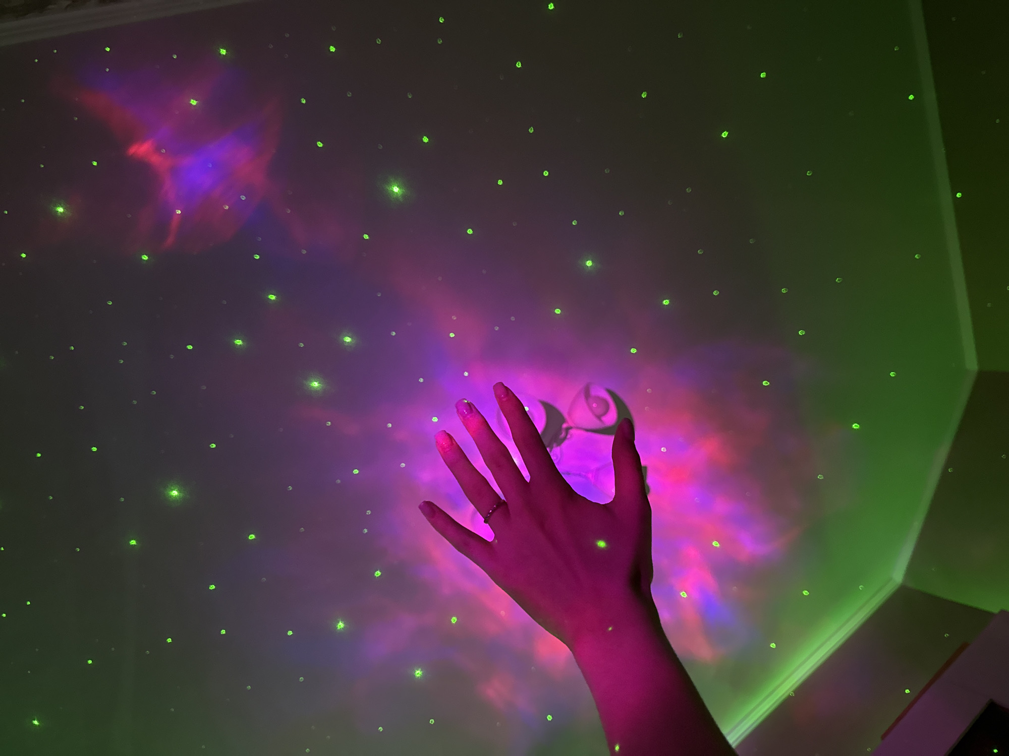 In an indoor setting, a person's left hand, illuminated by pink light and adorned with a silver ring, is extended towards the ceiling. The ceiling is projected with a colorful nebula-like pattern of pink and purple, interspersed with small green dots resembling stars. The light from the projection creates a surreal and atmospheric environment. The room appears to be a bedroom or living space with a traditional ceiling and wall junction.