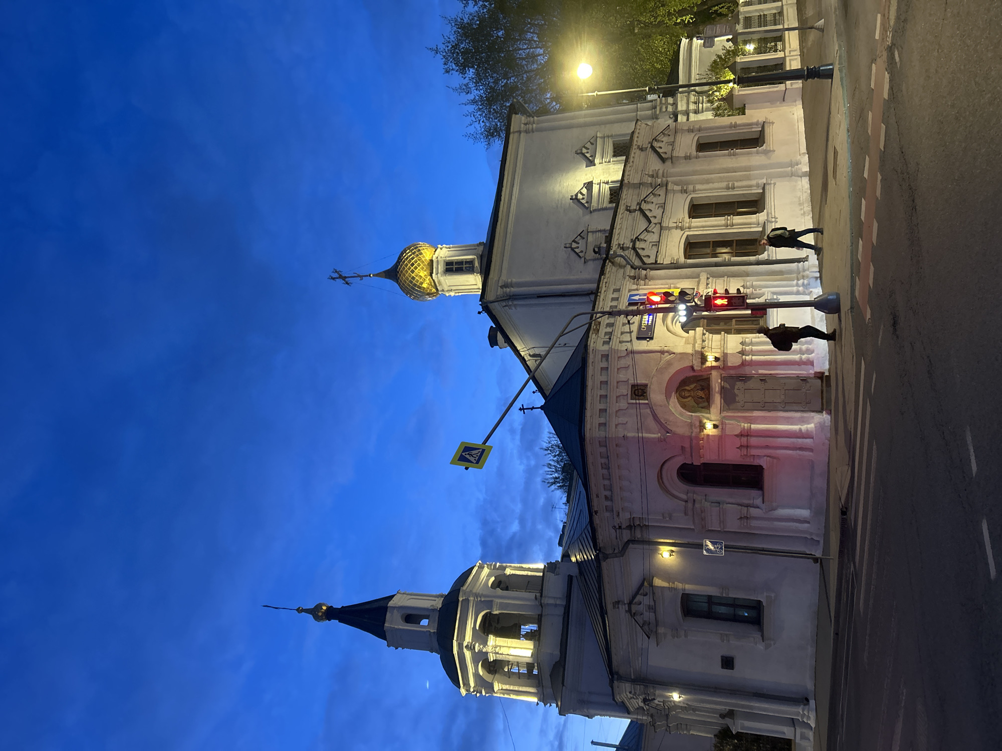 This evening image captures a street scene in Moscow, Russia, featuring historical architecture under a deep blue sky. The artificial lighting from streetlamps and illuminated buildings signifies dusk or early night.
Two prominent white buildings dominate the left side of the frame. On the far left, a tall, ornate bell tower rises, adorned with arched openings near its peak and capped with a dark, conical roof and a golden spire. To its right, another white building, characteristic of Russian Orthodox architecture, features intricate facades and is topped with a glistening golden onion-shaped dome surmounted by a cross. Both structures exhibit decorative elements around their windows and cornices.
In the foreground and extending towards the right, a paved road is visible, marked with white dashed lines and a solid red line. A prominent traffic light pole stands near the center, displaying red lights for vehicular traffic and a red "Don't Walk" symbol for pedestrians. On the control box attached to the traffic light pole, the Cyrillic text "СВЕТОФОР" (Svetofor), meaning "traffic light," is clearly visible.
Two individuals are present on the street: one person stands near the traffic light pole, and another is seen walking further down the road. A yellow diamond-shaped pedestrian crossing sign is mounted on a pole, extending from the bell tower building. A street lamp on the right casts a warm light on nearby tree foliage and parts of the building, contributing to the tranquil urban atmosphere.