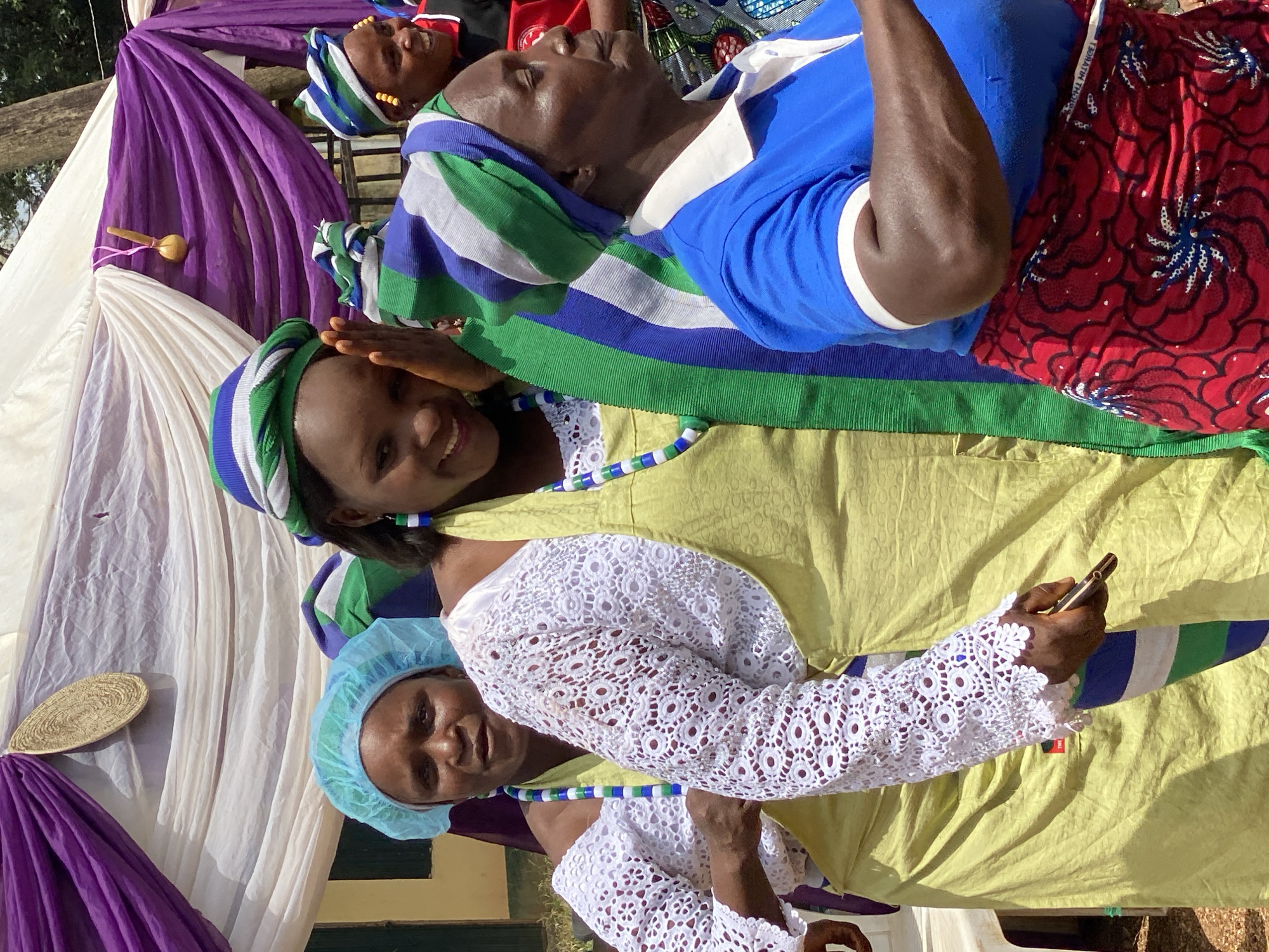 The image shows a group of women in Maigizo, Nigeria. One woman in the foreground smiles and shields her eyes. She wears a striped head covering with green, blue, and white, a yellow top, and a white lace sleeve. A woman wearing a light blue hair covering is partially visible below her. Another woman in the background wears a similar striped head covering with a blue shirt. A red patterned cloth is also in view. The setting is outdoors, with white and purple draping visible in the background. A small musical instrument hangs on the draping. A woven item is also attached to the cloth.