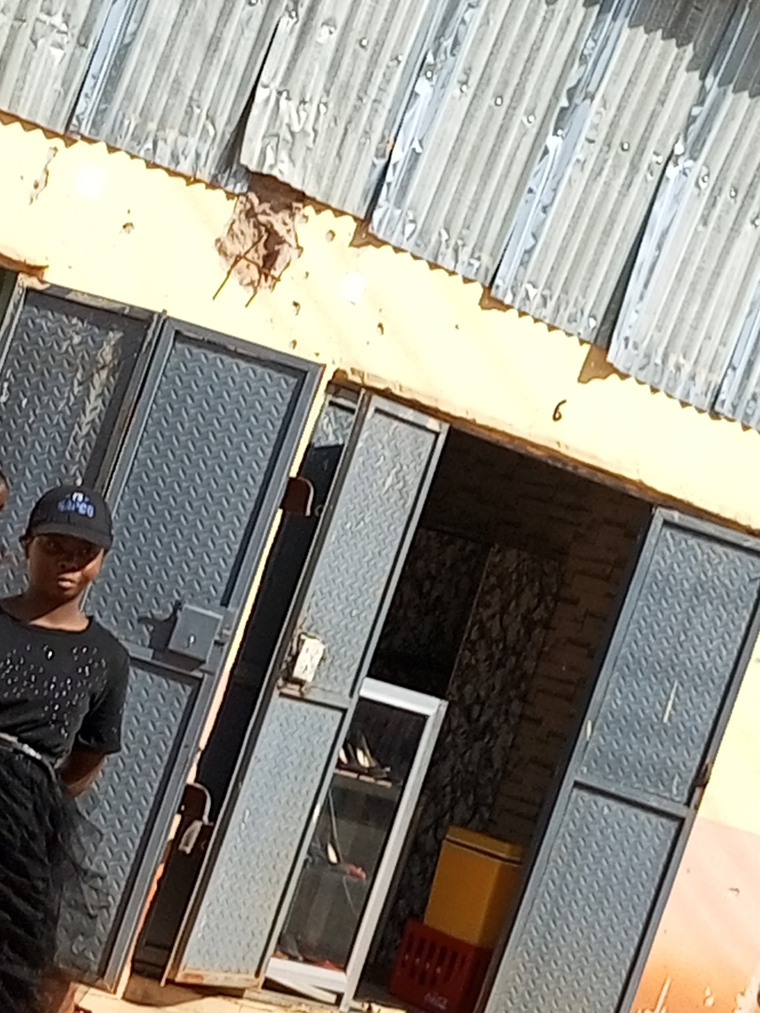 The image shows a young woman wearing a black t-shirt and a black baseball cap, standing in front of a building. The building has a corrugated metal roof and a yellow facade. There are two large metal doors on the building, one of which is open, revealing a dark interior. Inside the building, some items are visible, including what appears to be a glass cabinet and a red crate.
The woman is looking towards the camera, and her expression is neutral. Her t-shirt is decorated with white sparkling embellishments. She is wearing a black skirt. The scene appears to be outdoors, and the lighting suggests it is daytime. The presence of the building and the woman indicates a potentially urban or semi-urban setting. There are no clear indicators of specific weather conditions, but the bright lighting suggests sunny conditions.
A small, isolated "6" is visible on the yellow wall of the building, above and to the right of the open doorway. No other text is clearly legible in the image, though the cap the woman is wearing appears to have some writing on it, but it is not readable. The overall impression is of a casual, everyday scene. The context provided is Gombe, Nigeria.