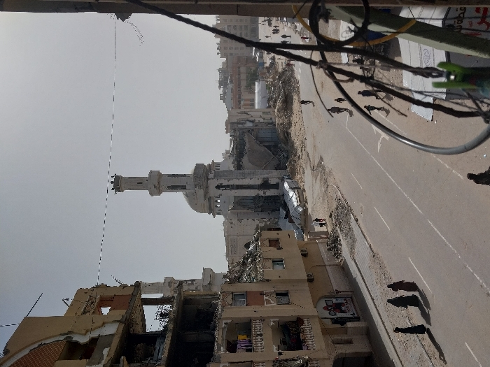 An elevated view captures a scene of widespread urban destruction in Khan Yunis, Palestinian Territory, under a hazy sky. In the mid-ground, a prominent white mosque with a domed roof and a tall, slender minaret stands significantly damaged, with large sections of its structure collapsed and interior elements exposed. The minaret itself shows damage at its apex. To the left, a multi-story residential building is heavily compromised, its facade partially destroyed, revealing exposed rebar and internal rooms. Clothes are visible hanging on balconies of the less damaged lower floors of this building. Debris, rubble, and dirt are extensively scattered across the ground around the structures.
A paved road bisects the scene, showing faint lane markings and accumulations of dirt on its verges. Several individuals are present on the street and amidst the debris. On the left side of the road, three women dressed in long, dark garments walk from left to right. Further down the street on the right side, multiple smaller figures, including adults and possibly children, walk in various directions. Near the base of the damaged mosque, several figures navigate the rubble-strewn ground. In the immediate foreground on the right, a blurred dark structure, possibly part of a building's exterior or utility pole with hanging wires and a green clothespin, partially obstructs the view. The scene depicts an active streetscape within a severely damaged urban environment.