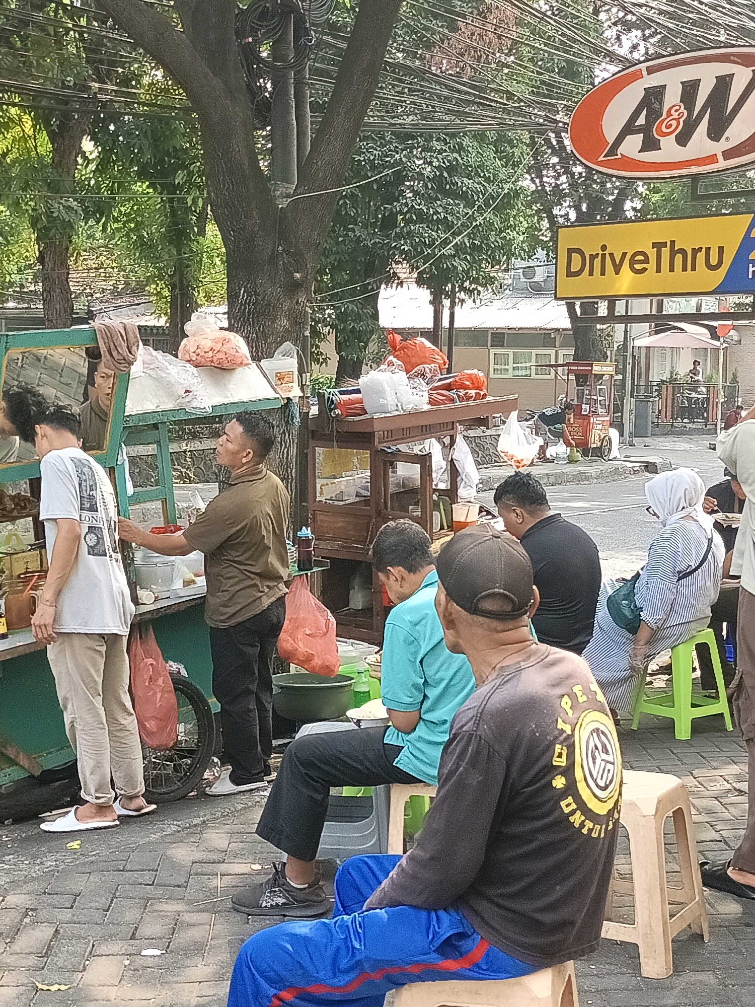 The image depicts an outdoor scene in front of an A&W restaurant on a sunny day, likely in South Jakarta, Indonesia, as indicated by the signage. In the foreground, several individuals are seated on plastic stools. A man in a black long-sleeved shirt with a circular yellow emblem on the back and blue track pants is seated on a beige stool. To his left, a man in a light blue t-shirt and dark pants is also seated on a stool. Further to the left, a woman wearing a white hijab and a striped top is seated on a bright green stool, appearing to interact with someone behind her.
To the left of the seated patrons, a food stall with a green frame is visible. A man in a brown short-sleeved shirt is attending to the stall, interacting with a customer or employee. This individual is wearing dark pants. Another person, wearing a white t-shirt and light-colored pants, stands at the stall, facing away from the camera. The stall displays various items, including bags of snacks and drinks. Red plastic bags are hanging from the stall's structure.
In the background, a yellow sign with the text "DriveThru" is prominently displayed, along with the A&W logo. Trees with lush green foliage line the area, and power lines are visible overhead. The ground is paved with brick-like tiles. Further in the background, more structures and a glimpse of parked vehicles are present, suggesting a commercial or urban environment. The overall atmosphere appears to be that of a casual dining or street food setting.