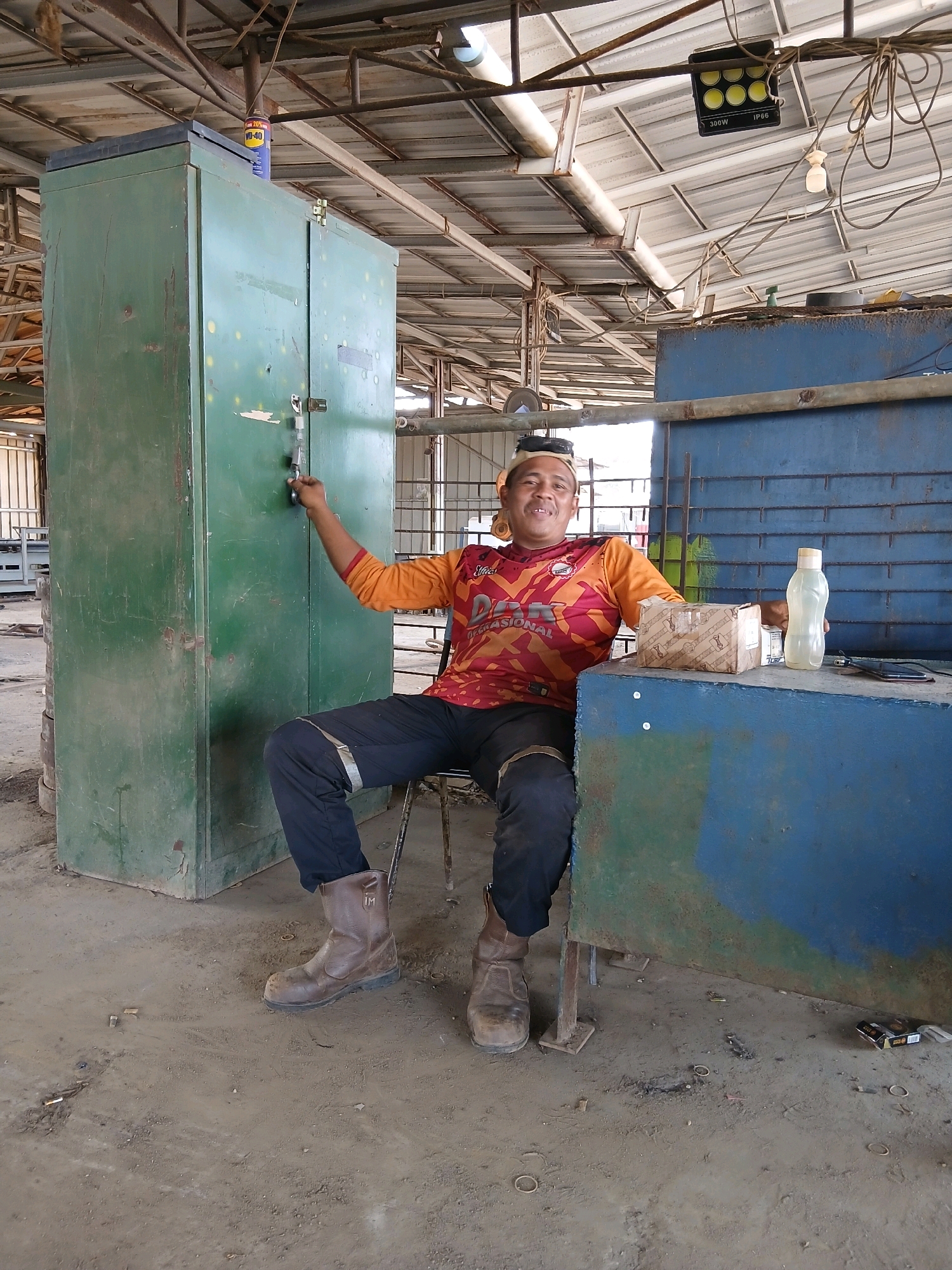 A man in an orange and red long-sleeved shirt and black pants with reflective stripes is sitting on a chair in front of a blue structure in what appears to be an industrial or workshop setting in Pangkalpinang, Indonesia. He is wearing brown work boots. His right hand is extended towards a large, green metal cabinet, which has a latch that is partially open. A key is visible in the lock mechanism. The man is smiling and looking towards the camera. On the blue structure in front of him, there is a cardboard box, a clear plastic water bottle, and a dark object, possibly a phone. The environment is dusty and unkempt, with a corrugated metal roof overhead supported by metal beams. Various items, including debris and discarded cigarette butts, are scattered on the ground. In the background, there are glimpses of other industrial structures.