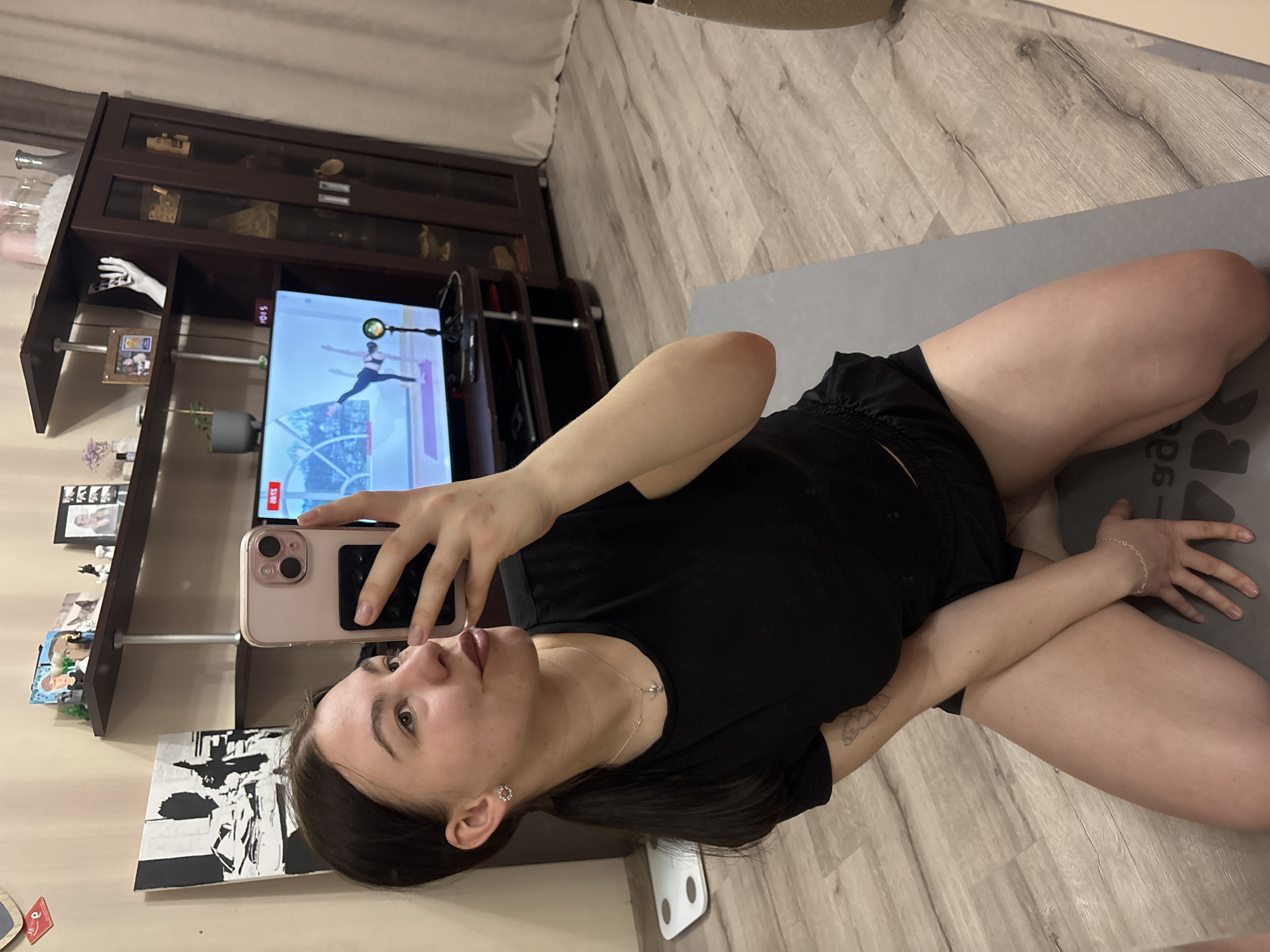 A woman is taking a selfie indoors. She is on a gray yoga mat on a light-colored wooden floor. She is wearing a black t-shirt and shorts. In the background, there is a dark wooden entertainment center with a TV showing a yoga workout. There is also a small shelf with various decorations and pictures. A weight scale is visible by the wall. The location is Perm, Russia, based on the metadata of similar images.