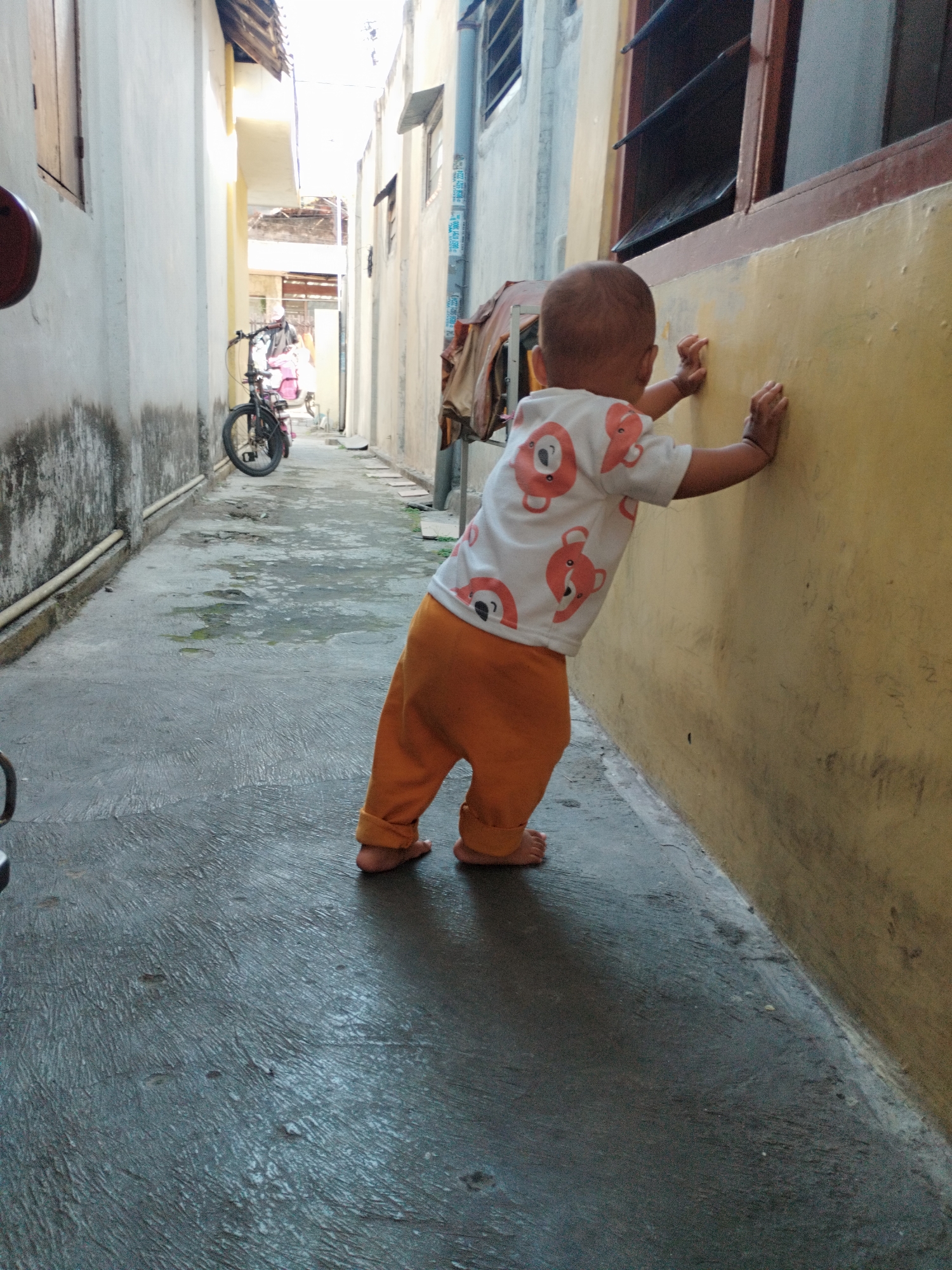 A toddler, wearing an orange bottom and a white shirt with a bear pattern, stands with their hands against a yellow wall in an alleyway in Surabaya, Indonesia. The toddler appears to be learning to walk or exploring their surroundings. The alleyway is narrow, with concrete walls on either side. There is a bicycle parked in the background. The ground is wet and reflective, suggesting recent rain. The scene is captured in daylight.