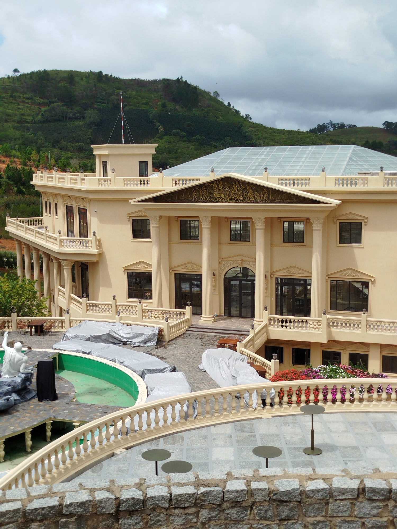 The image shows a large, cream-colored building with classical architectural elements, including columns and ornate friezes, set against a backdrop of lush green hills. The building features multiple levels, balconies, and a glass-paneled roof. In the foreground, there is a stone wall and a balcony with a railing, overlooking a patio area with two small tables and chairs. Below the balcony, a semicircular swimming pool with a green lining is partially covered with grey tarps, and a white statue of a seated figure is visible. The overall scene suggests a resort or a large private residence in a hilly, possibly tropical, environment. The location is Lạc Dương, Vietnam.