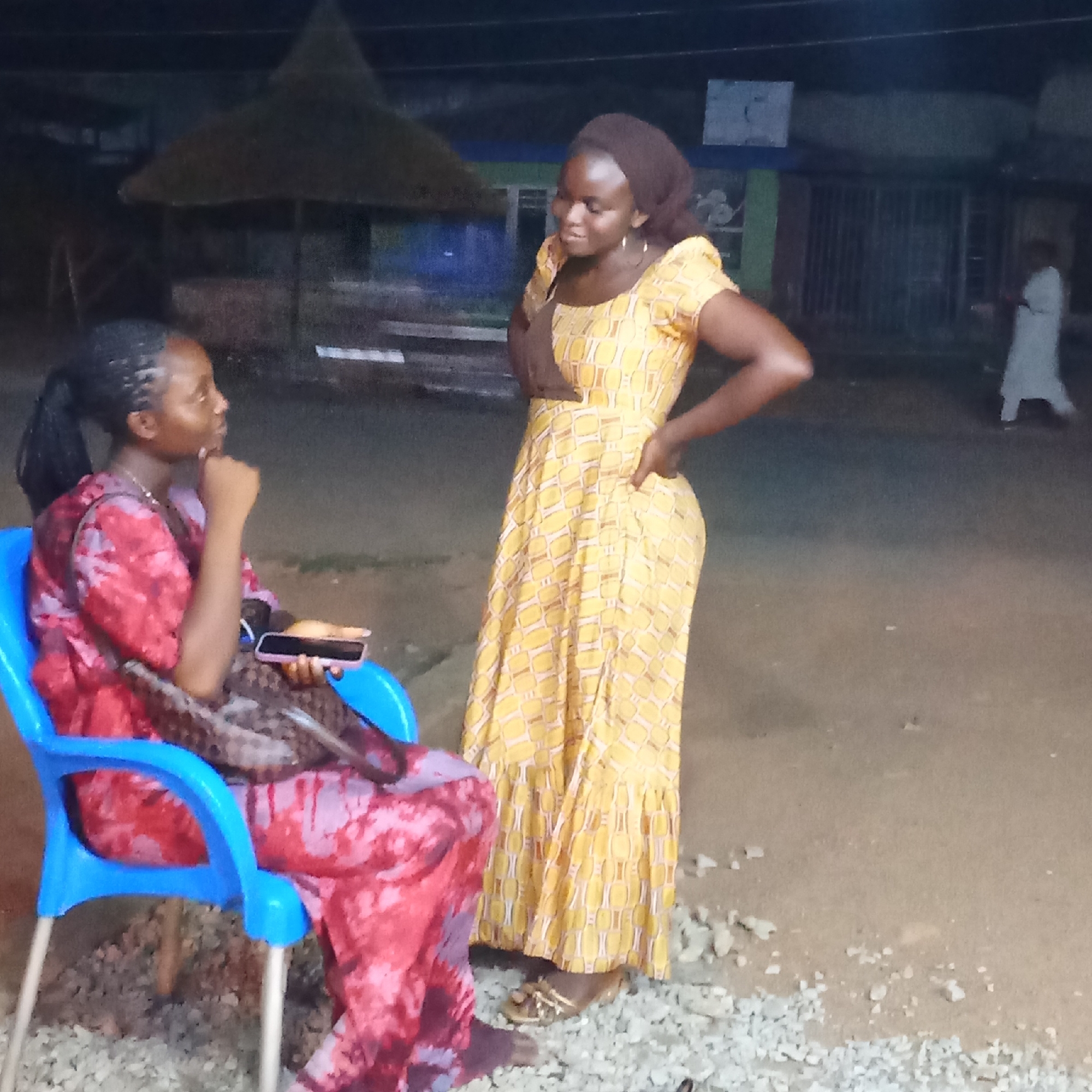 The image shows two women standing in an outdoor setting in Akwanga, Nigeria, at night. One woman is seated on a blue plastic chair, wearing a tie-dye dress and holding a cell phone. The other woman stands next to her, wearing a yellow patterned dress and a brown head covering. The ground is covered in small rocks. Behind the women is a thatched-roof structure. A person wearing a white garment is walking in the background. There is a building behind the women.