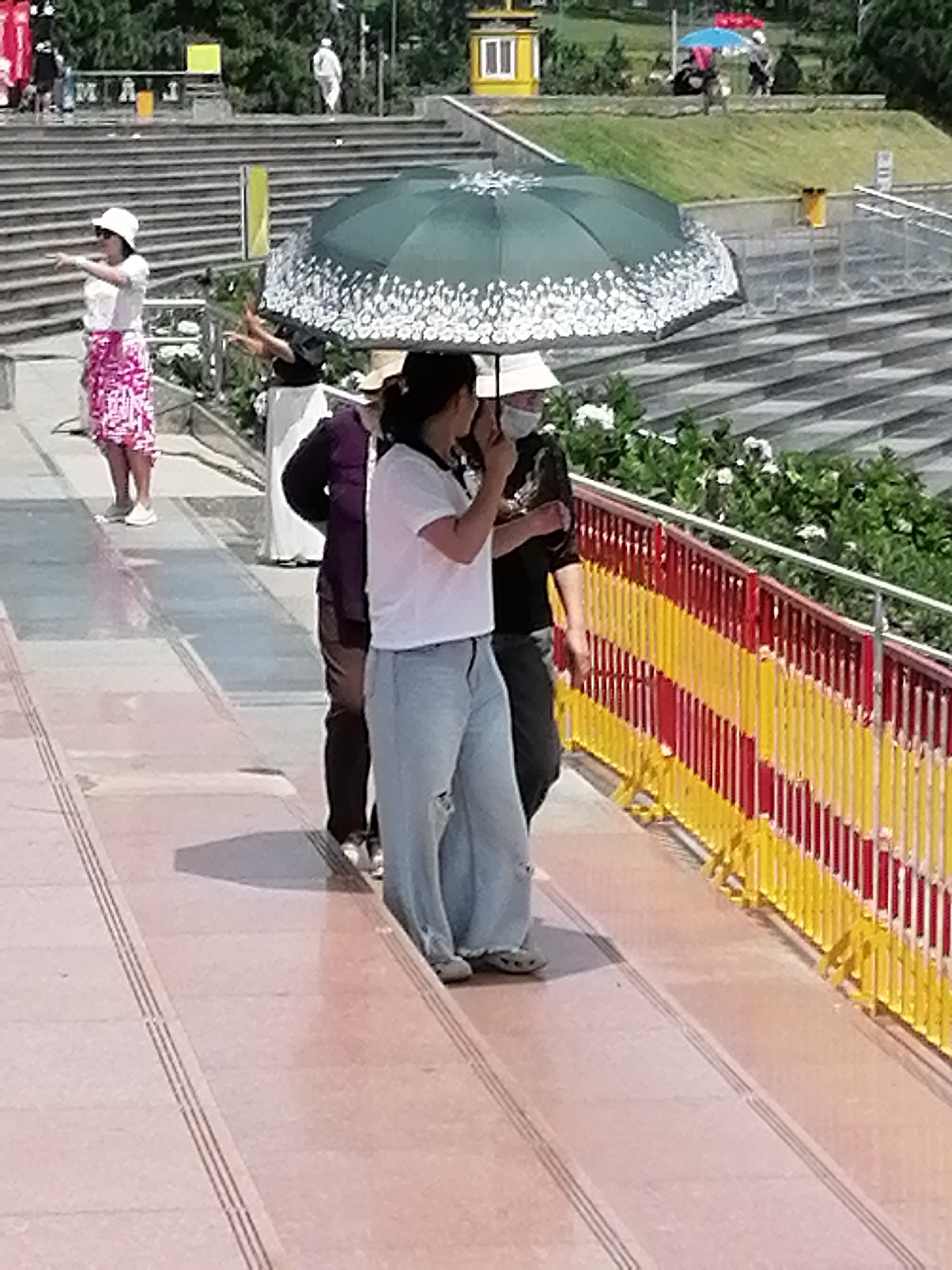 In the city of Da Lat, Vietnam, a group of individuals are gathered on a wide, paved walkway with a reddish-pink hue, bordered by a yellow and red striped railing. The walkway is marked with dark parallel lines. Behind the walkway, tiered stone stairs ascend a grassy slope, and further back, lush green trees are visible. On the walkway, a woman wearing a white hat and a white top with a pink and white patterned skirt stands facing left, with her right arm extended, possibly pointing. To her right, a person in a purple top and dark pants is partially obscured. In the center of the image, two individuals are walking under a large, dark green umbrella adorned with white floral patterns, shielding them from the sun. The person in front, on the right, is wearing a white t-shirt and light blue jeans. The other person, on the left and behind, is wearing a dark shirt. The scene suggests a sunny day, likely a tourist area, given the presence of umbrellas and the open-air setting.