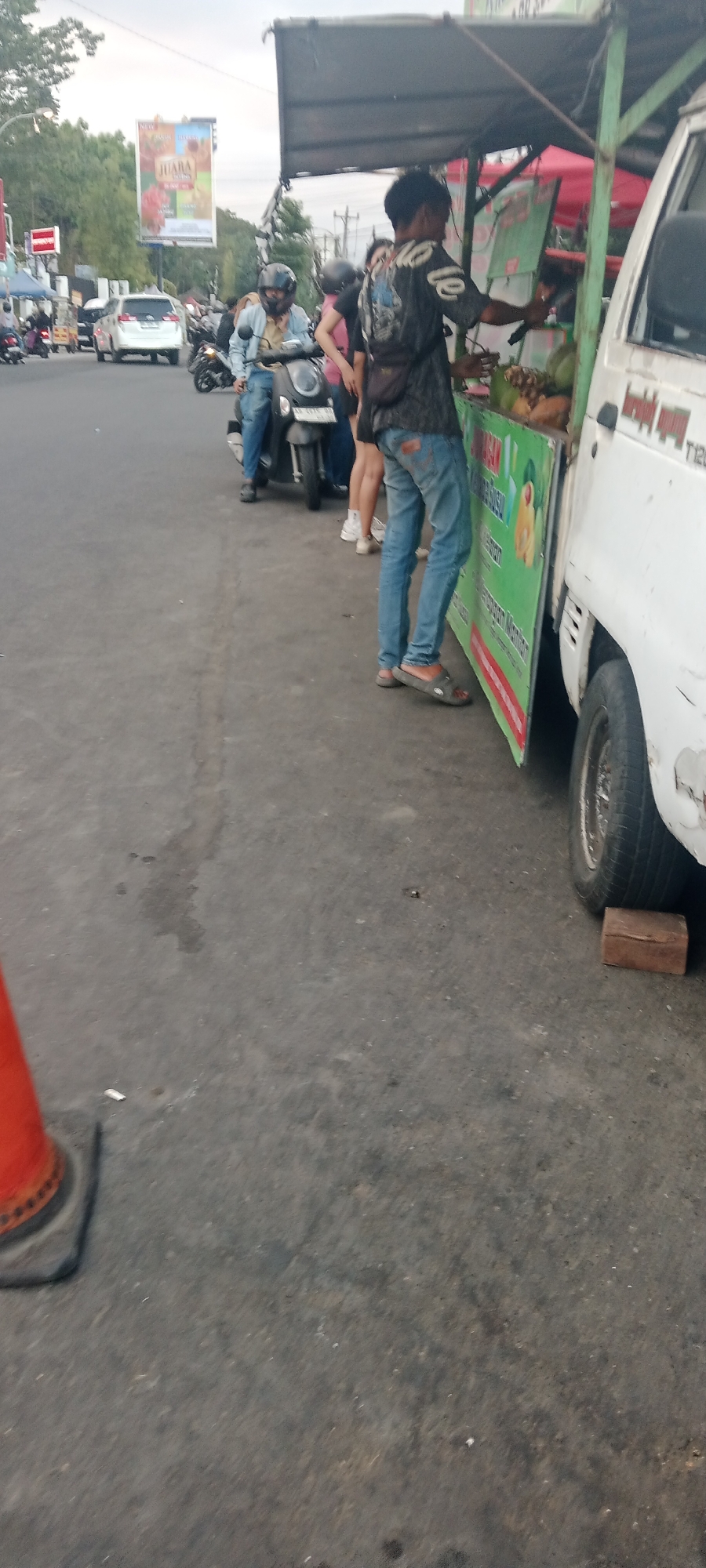 A street scene in Ngemplak, Indonesia, shows a roadside vendor selling coconuts from a cart attached to a white truck. A young man wearing a dark patterned shirt, blue jeans, and gray sandals stands at the counter, appearing to be a vendor or customer. He holds a small knife in his right hand. Behind him, two women are visible, one in a pink shirt and the other in a black top and jeans. A man on a scooter wearing a blue jacket and a helmet is seated to the left. In the background, there is a billboard with the word "JUARA" and images of flowers, traffic, and trees. The ground is asphalt, with an orange traffic cone and a piece of wood supporting the truck's wheel. The sky is overcast.