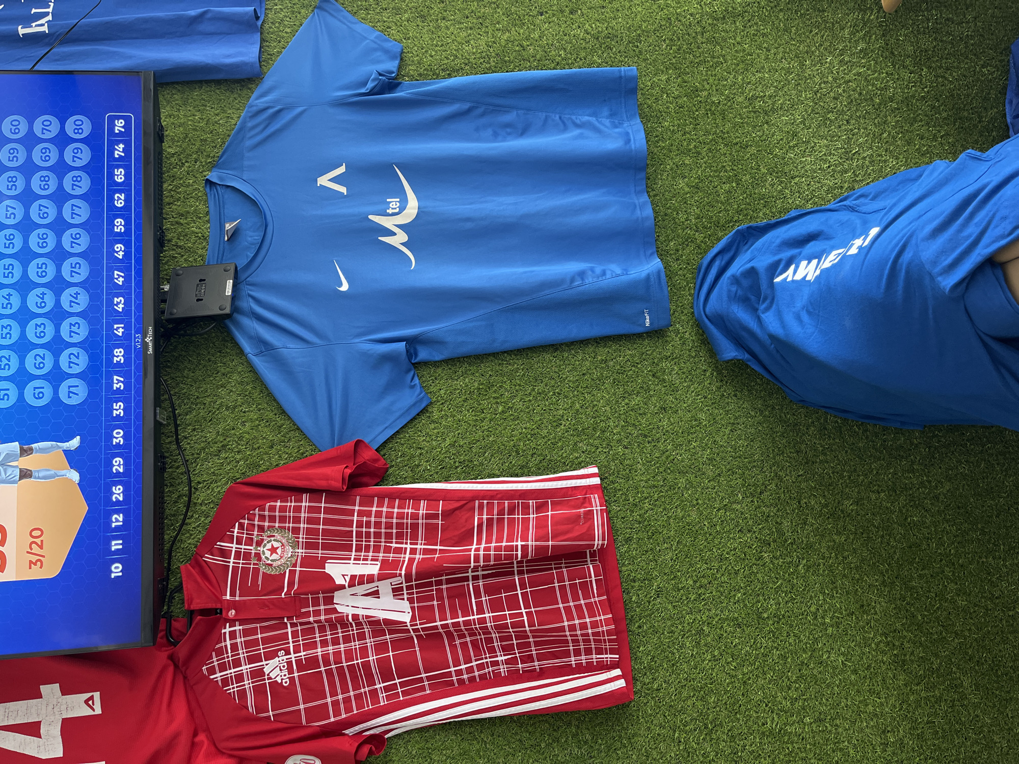 This overhead shot depicts a sports setting, likely indoors in Ivailo, Bulgaria, with objects laid out on a vibrant green artificial turf surface.
The main subjects are two soccer jerseys and a monitor screen. A blue Nike jersey is centrally placed, featuring a white Nike swoosh, the "M-tel" logo, and another white symbol resembling a caret or "V" on the chest. "NikeFIT" text is visible on its lower right. Another blue fabric item, possibly a part of the same kit, lies partially visible to the right with reversed white text, appearing to read "MY".
Below it is a red Adidas jersey, sporting a distinct crest with a star and a laurel wreath on the left chest, and a prominent white number "4" on the front. The "adidas" logo is visible, along with partially visible "CLIMAC" text (for Climacool technology). White stripes adorn the shoulders and sides of the jersey.
To the left, a monitor displays a grid of numbers from 10 to 80 within blue circular bubbles. Several numbers, including 76, 65, 62, 59, 49, 47, 43, 41, 38, 37, 35, 30, 29, 26, 12, 11, and 10, are highlighted with a white background, suggesting a selection or lottery process. A graphic of players is shown on the left of the screen, beneath a "3/20" indicator. A black device labeled "SMARTTECH V1.2.3" and "CERCO" is attached to the monitor's side. The scene implies a sporting event's preparation, perhaps a team lineup reveal or a raffle.
