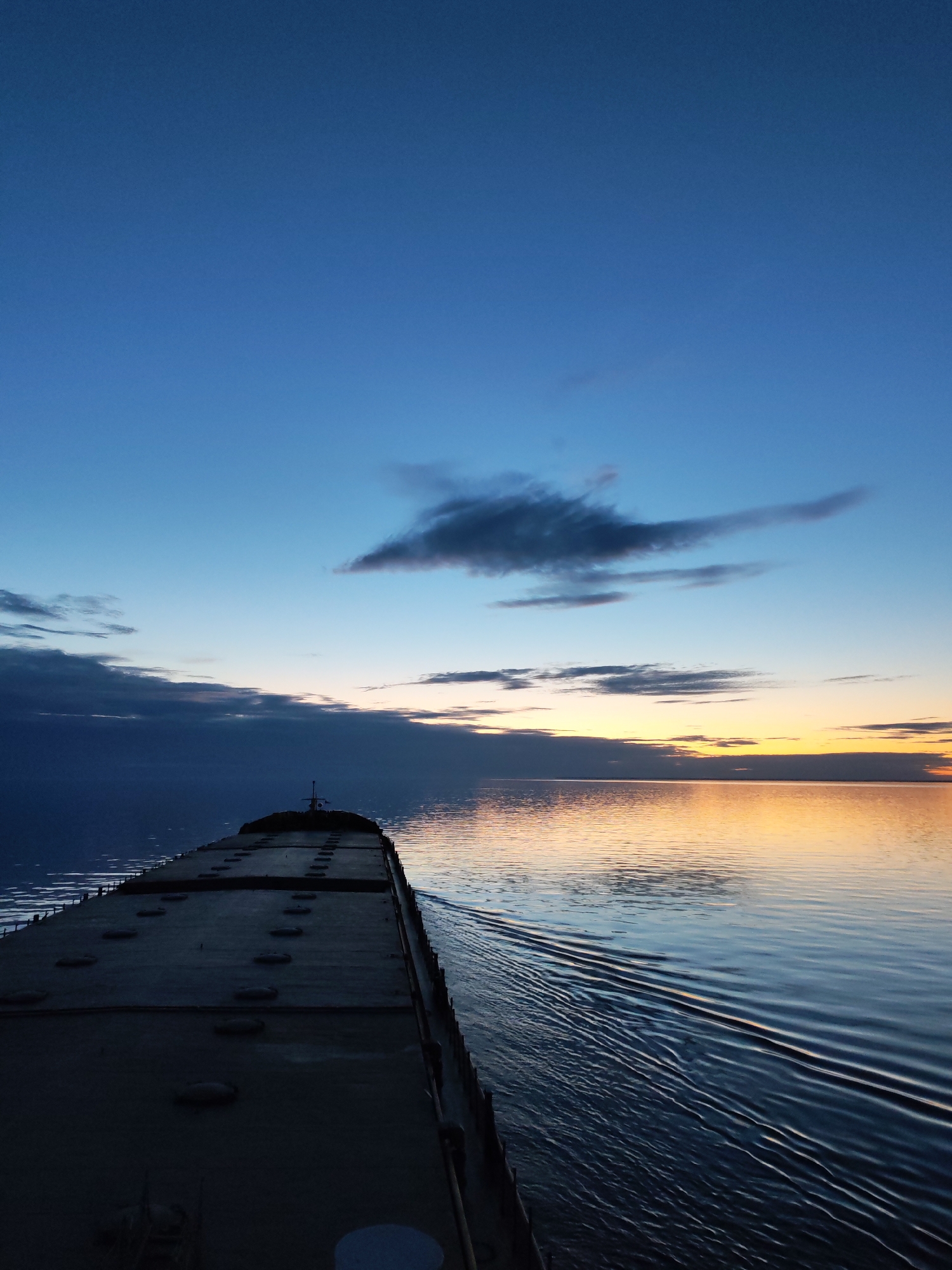 The image depicts a serene twilight scene aboard a large vessel traversing a vast body of water. The primary subject is the forward section of a long, flat-decked ship, possibly a bulk carrier, extending from the lower left towards the center of the frame. Its deck is dark, featuring multiple rows of circular, raised hatches. A small mast or structure is visible near the bow.
The vessel is in motion, evidenced by the distinct wake and parallel ripples fanning out across the calm water surface to its starboard side. These ripples beautifully reflect the warm colors of the horizon.
The sky exhibits a dramatic gradient, transitioning from a deep blue overhead to lighter shades of blue and then vibrant oranges, yellows, and pale pinks along the horizon. This illumination strongly suggests either sunrise or sunset. Scattered clouds are present, including a notable elongated, dark cloud formation in the upper middle of the sky, with smaller, wispy clouds near the horizon. The horizon line is clear, separating the tranquil water from the expansive sky. The overall atmosphere is one of peaceful transit amidst a beautiful natural light display. No people or text are visible.