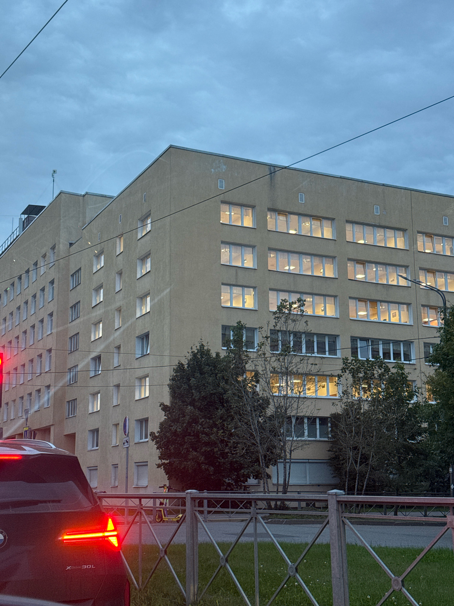 The image shows a large, light-beige, multi-story building with many windows, some of which are illuminated from within. The building has a modern, angular architectural style. In the foreground, the rear of a dark-colored SUV is visible, with its red taillights illuminated. A metal fence with a diamond pattern runs horizontally across the lower part of the image. Beyond the fence, there is a grassy area and a street. A yellow scooter is parked near a street sign. Trees and branches partially obscure the lower floors of the building. The sky is overcast and appears to be twilight. The scene suggests an urban setting, possibly a business or residential complex.