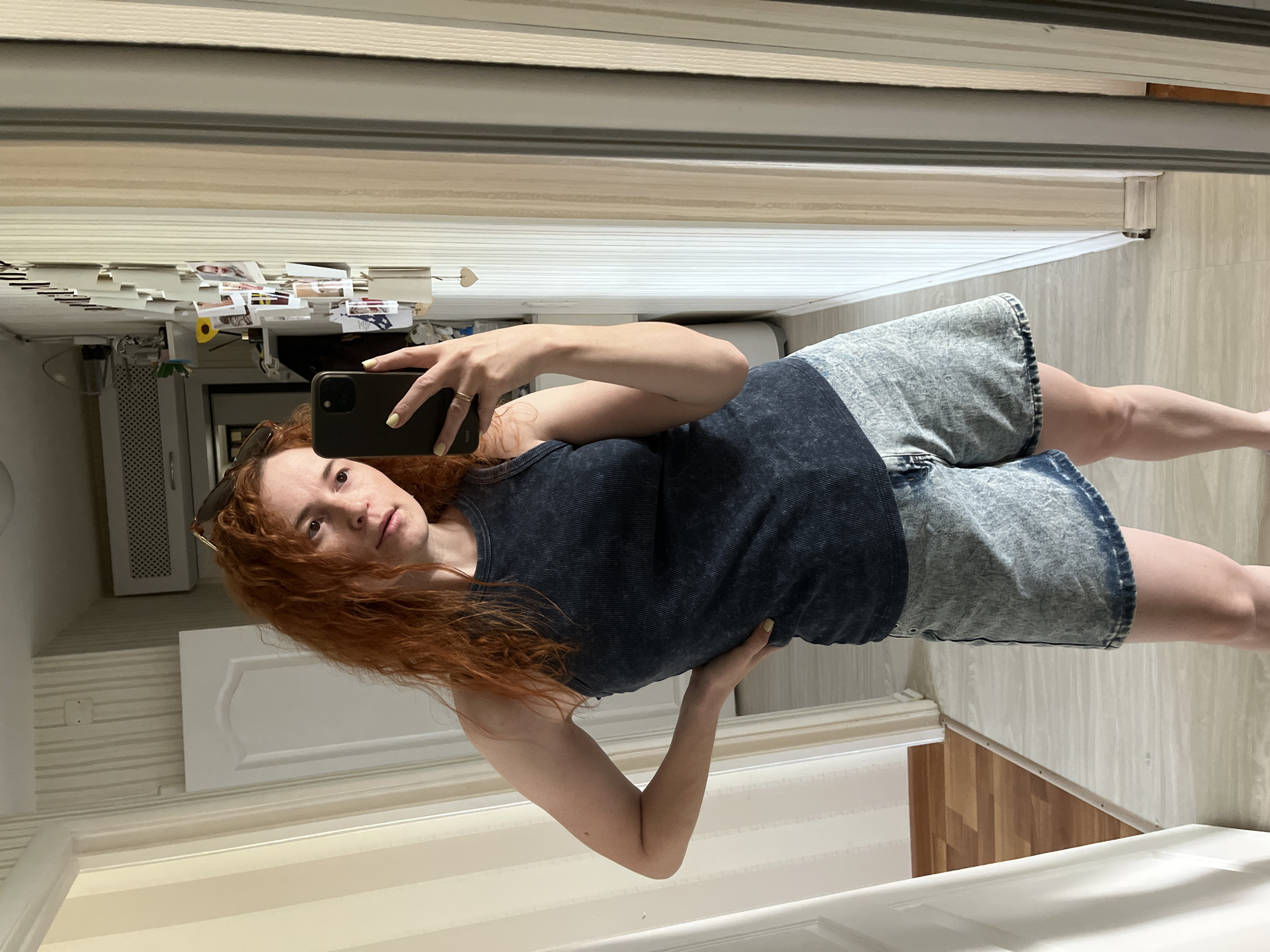 A red-haired woman takes a selfie in a mirror. She wears a dark blue tank top and acid wash shorts. The background reflects a hallway with light-colored striped wallpaper, a white door, and wood-like flooring. In the mirror's reflection, above the woman, are photos and papers attached to what appears to be a cabinet. The location is a domestic interior, potentially in the city of Chaikovsky, Russia.